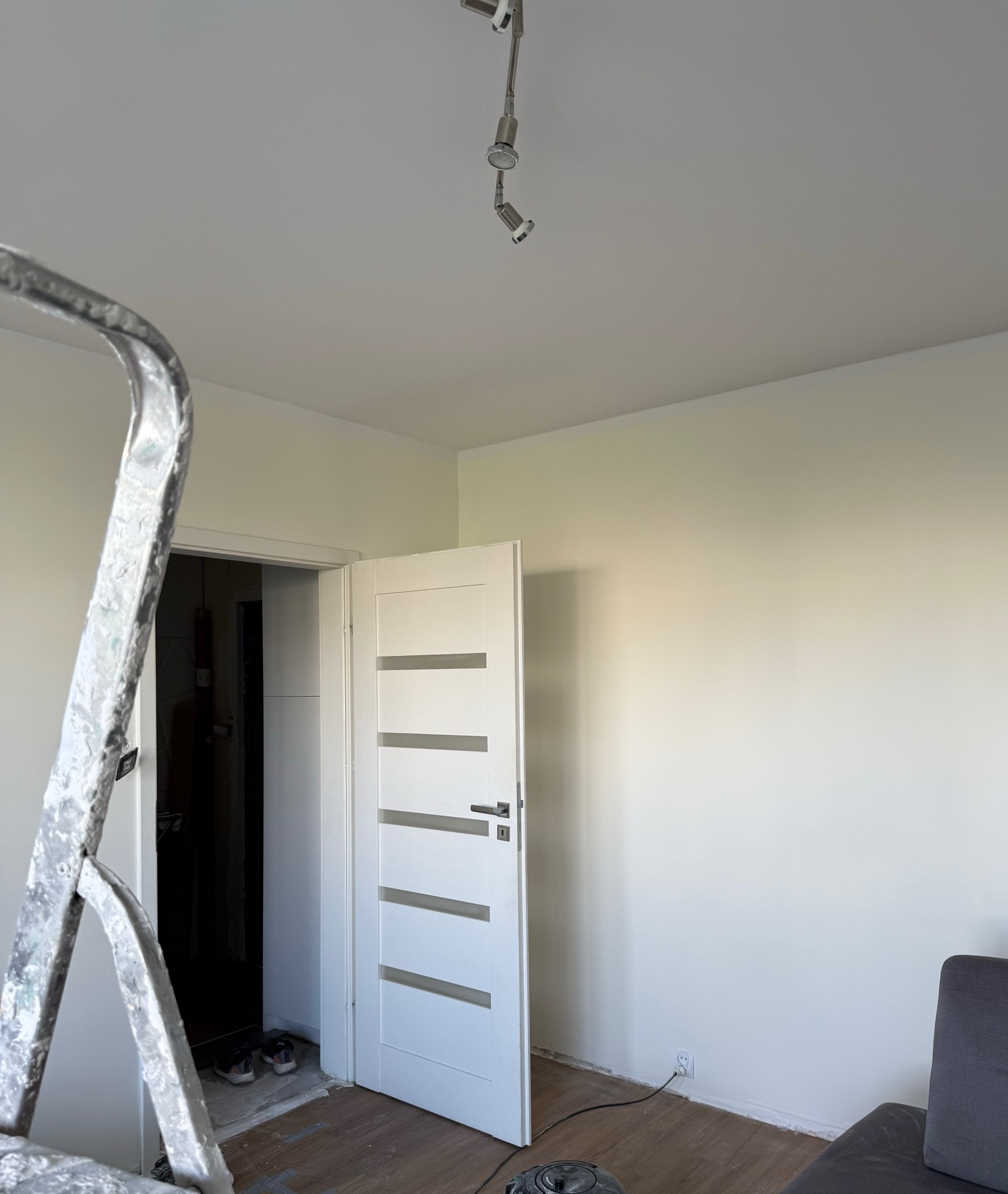 Wnętrze po malowaniu ścian na jasny odcień, widoczna drabina malarska, białe drzwi z przeszkleniami i fragment sofy. Parkiet na podłodze.