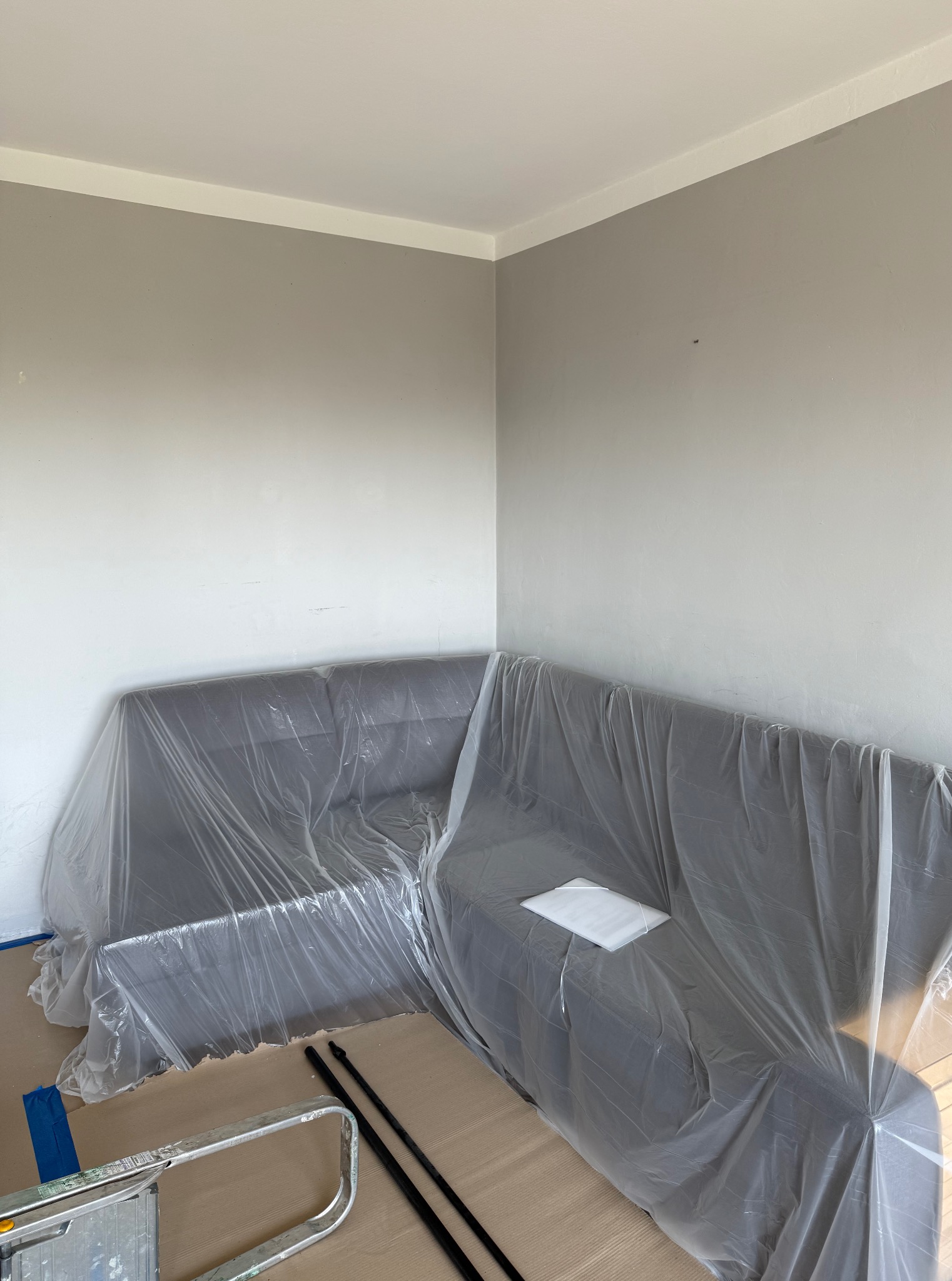 Pokój przygotowany do malowania: narożnik przykryty folią ochronną, widoczne drabina i wałki malarskie. Ściany w odcieniach szarości, listwa sufitowa biała.