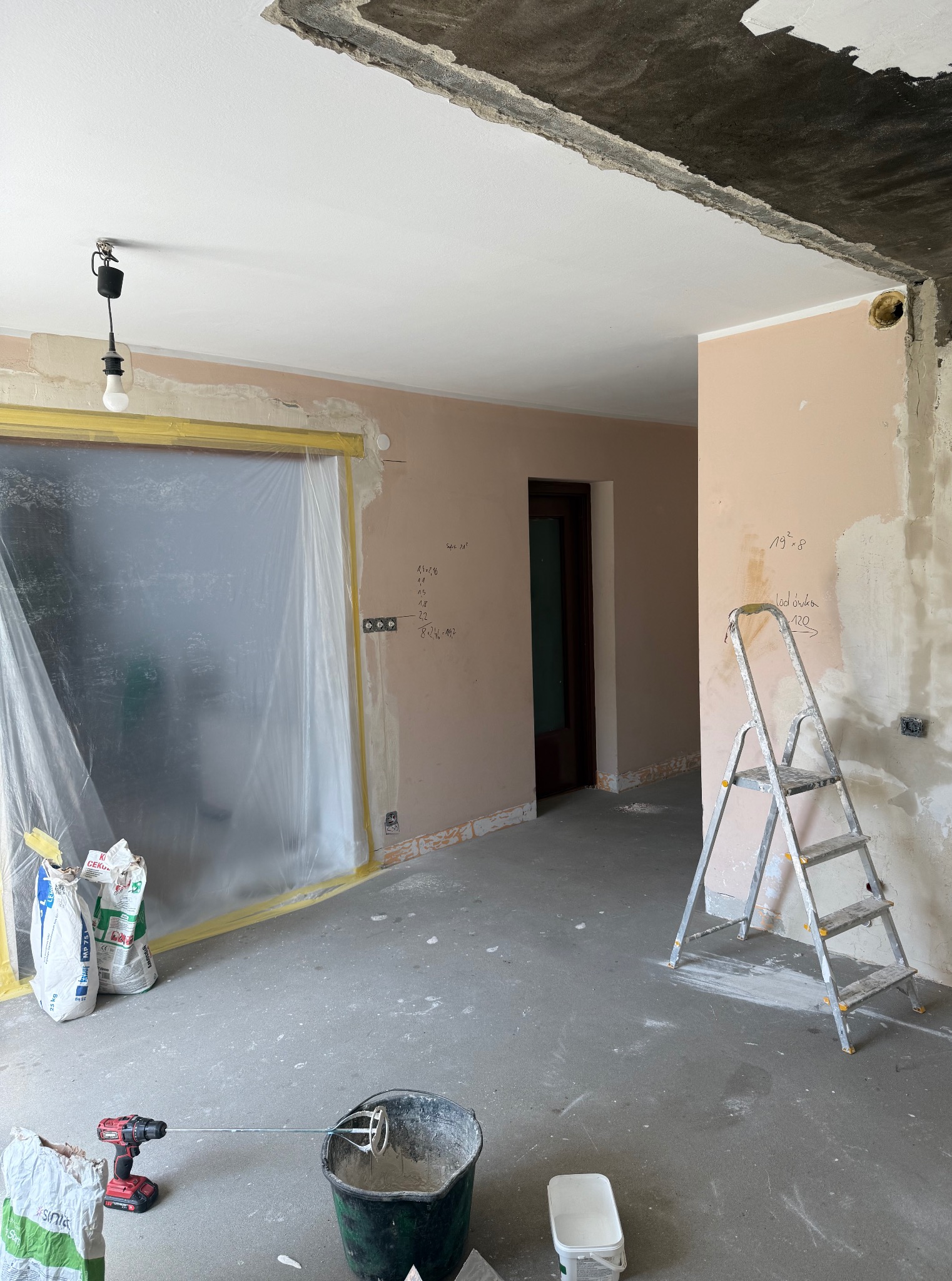 Wnętrze w trakcie prac: ściany przygotowane do malowania, zakryte folią okno, drabina, wiadro z zaprawą i mieszadło. Widoczne niedoskonałości sufitu.