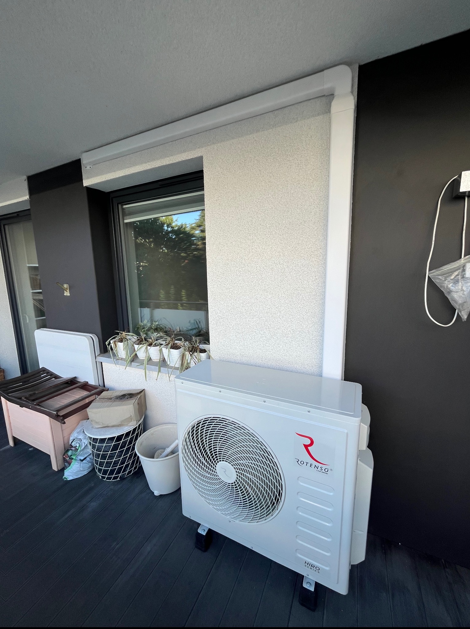 Biała jednostka zewnętrzna klimatyzacji Rotenso Hiro Series na balkonie z czarną podłogą i ścianą. Widoczne okno z roślinami, elementy wyposażenia balkonu.