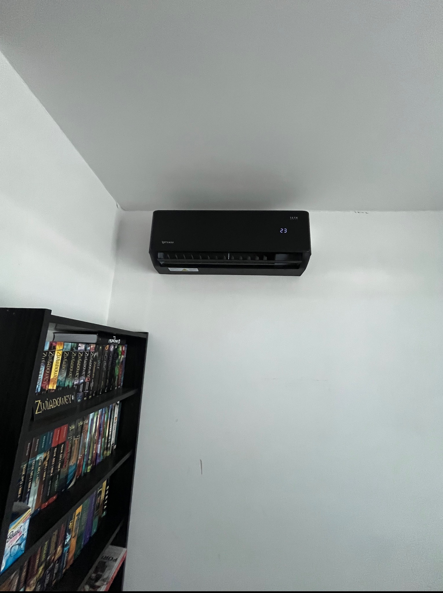 Czarna klimatyzacja Rotenso TATA 23 zamontowana na białej ścianie, obok czarnego regału z książkami. Widok z dołu, oświetlenie dzienne.
