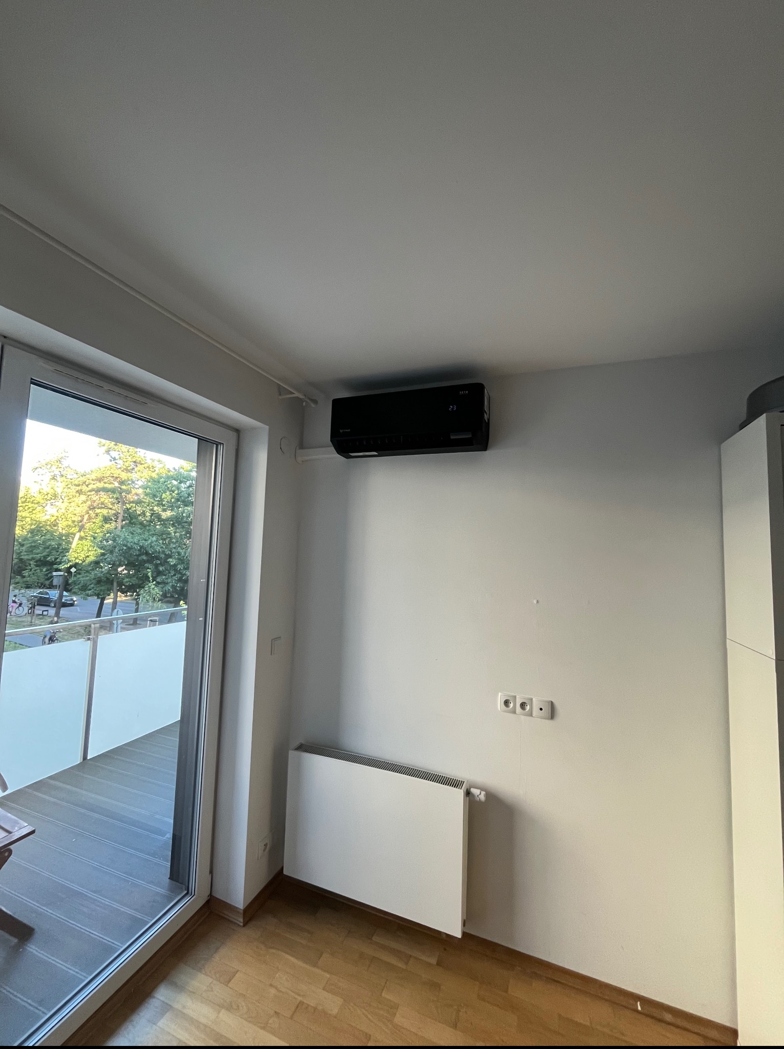 Czarny klimatyzator ścienny w jasnym pokoju z balkonem. Widoczny grzejnik panelowy i drewniana podłoga. Minimalistyczny design.