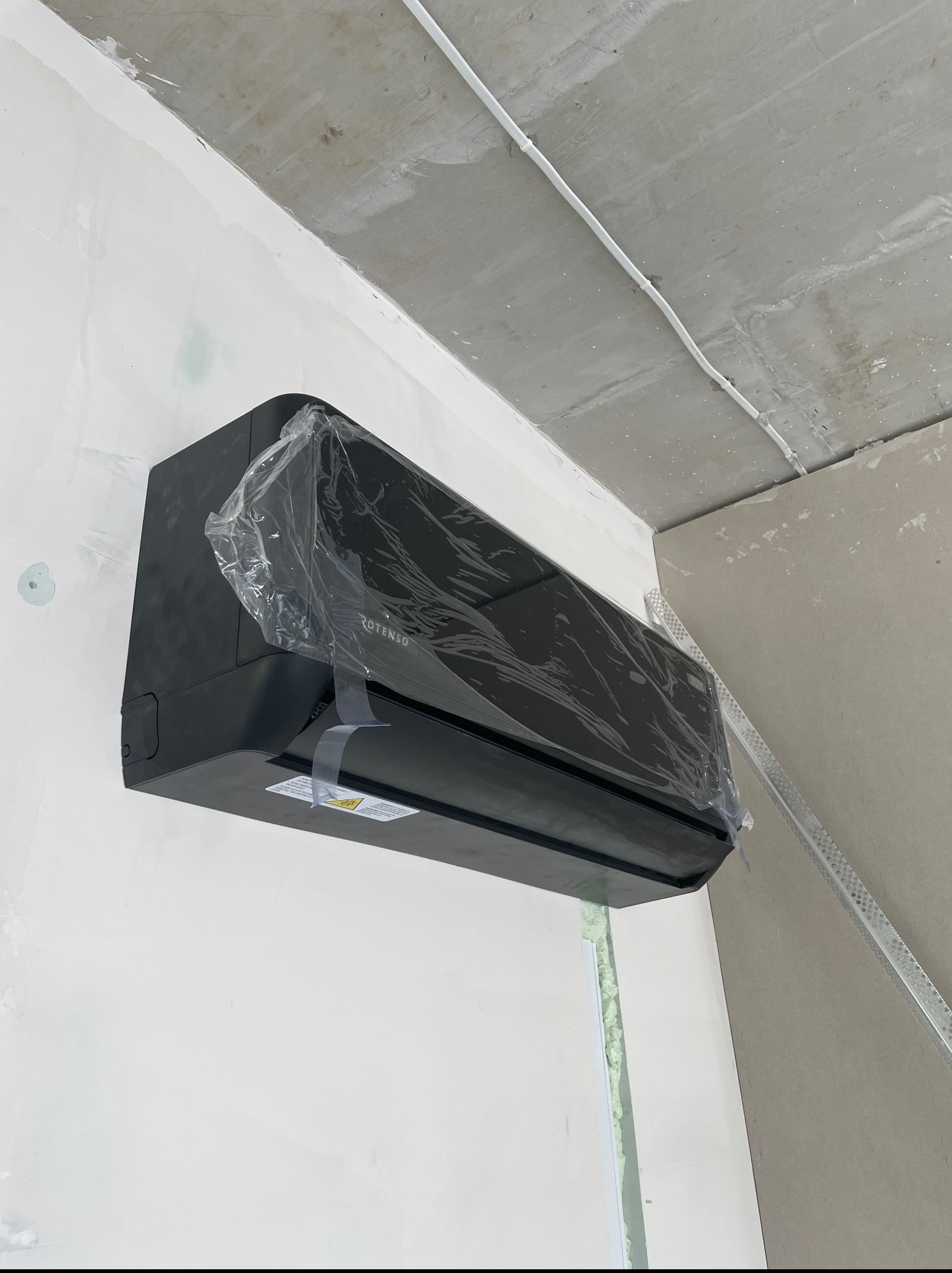 Czarna jednostka wewnętrzna klimatyzacji Rotenso, jeszcze w folii ochronnej, zamontowana na ścianie w trakcie prac wykończeniowych. Widoczne kable i szpachlowane ściany.