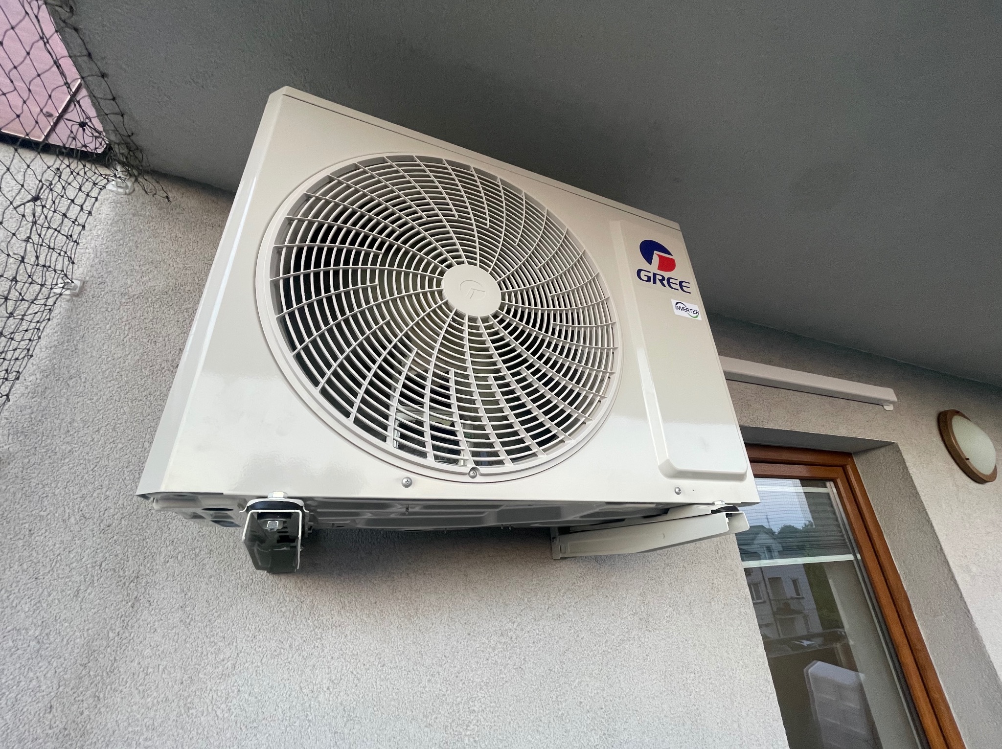 Zamontowana jednostka zewnętrzna klimatyzacji Gree na balkonie, widok z dołu. Widoczne okno i siatka zabezpieczająca na tle szarej ściany budynku.
