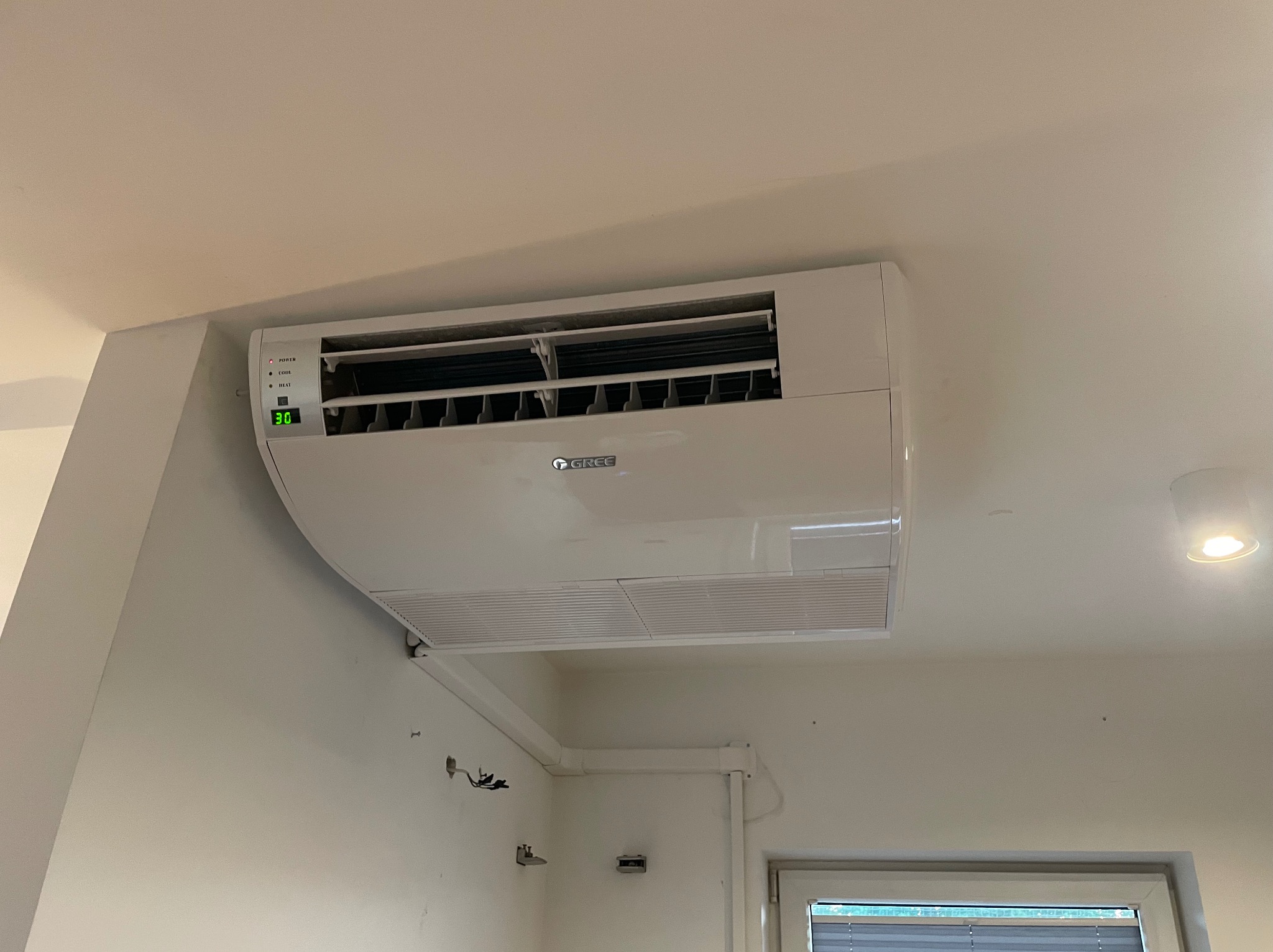 Biała klimatyzacja marki Gree zamontowana na ścianie w pomieszczeniu. Widoczne przewody i rury instalacyjne. Na wyświetlaczu klimatyzacji widoczna temperatura 30 stopni.