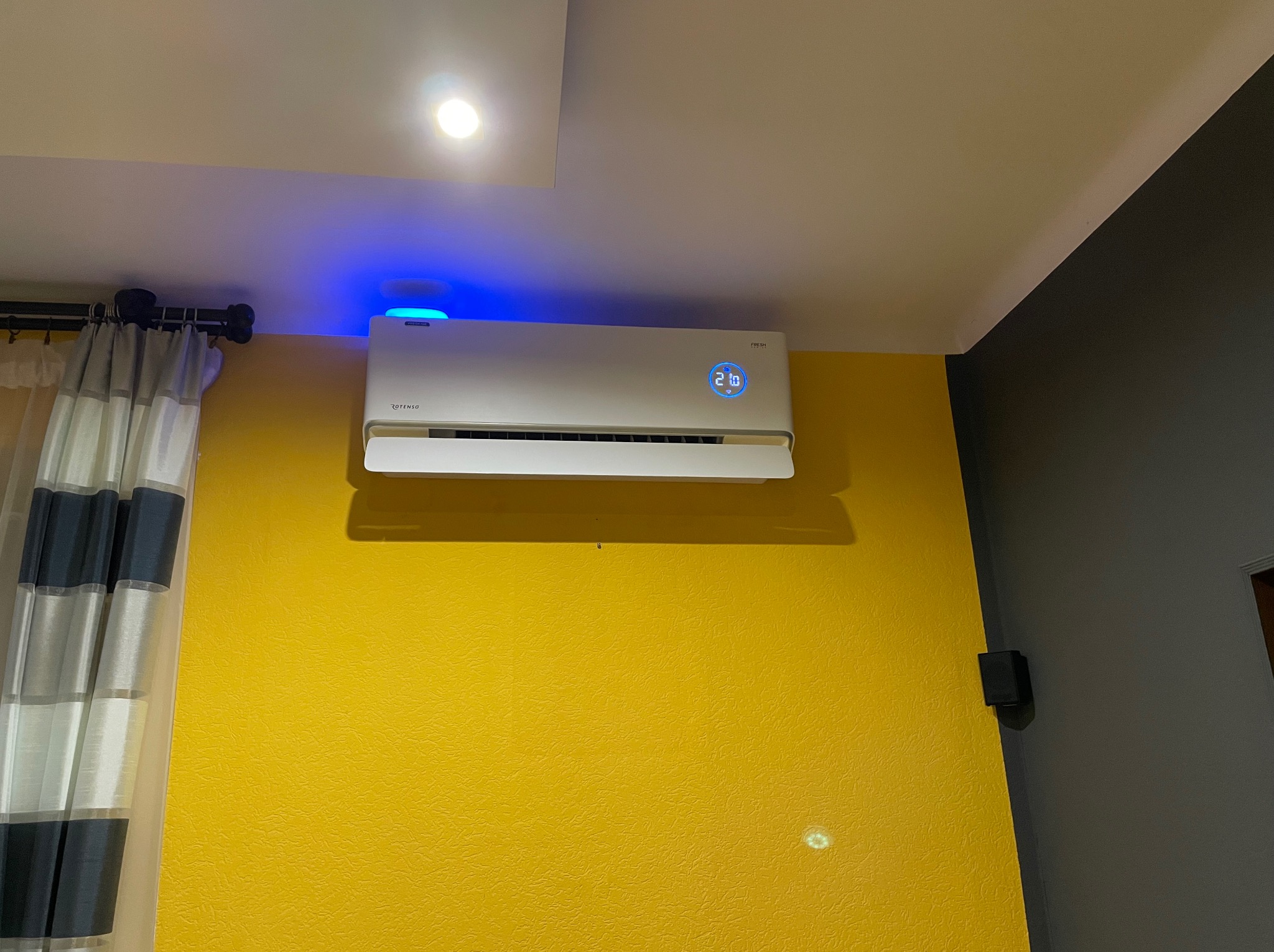 Biała klimatyzacja z niebieskim podświetleniem i wyświetlaczem 21 stopni Celsjusza na żółtej ścianie. Widoczne zasłony i fragment szarej ściany.