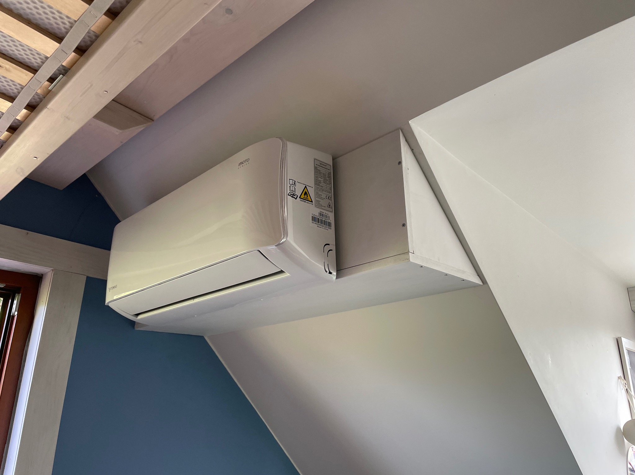 Biała klimatyzacja split marki IMOTO zamontowana pod skosem na poddaszu. Widoczne elementy konstrukcyjne dachu i fragment okna. Ściana pomalowana na dwa kolory.
