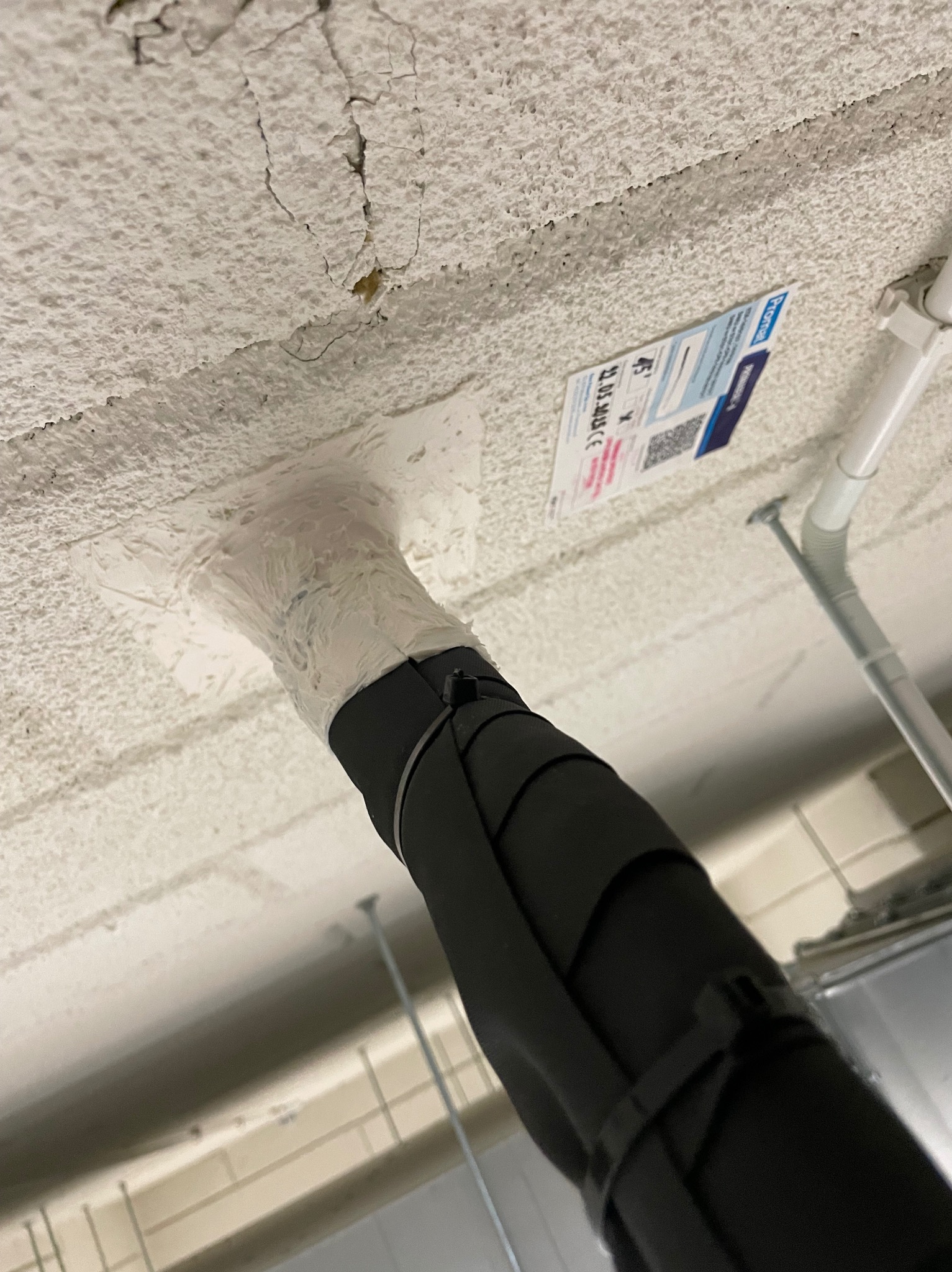 Czarny przewód klimatyzacji przymocowany do sufitu z widocznym uszczelnieniem i naklejką informacyjną. Widok z dołu, sufit z fakturą baranka.