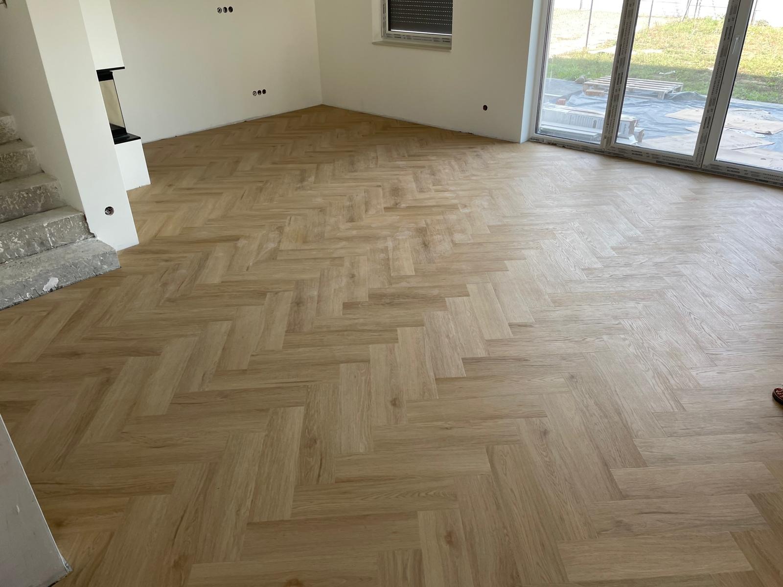 Jasny parkiet ułożony we wzór jodełki francuskiej w przestronnym, jasnym pokoju z oknem i surowymi schodami.