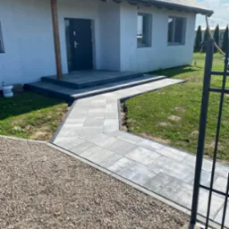 Ułożona kostka brukowa tworzy ścieżkę do domu z szarymi schodami. Obok ścieżki znajduje się obszar wysypany drobnym żwirem i trawnik. W tle widać fragment ogrodzenia.