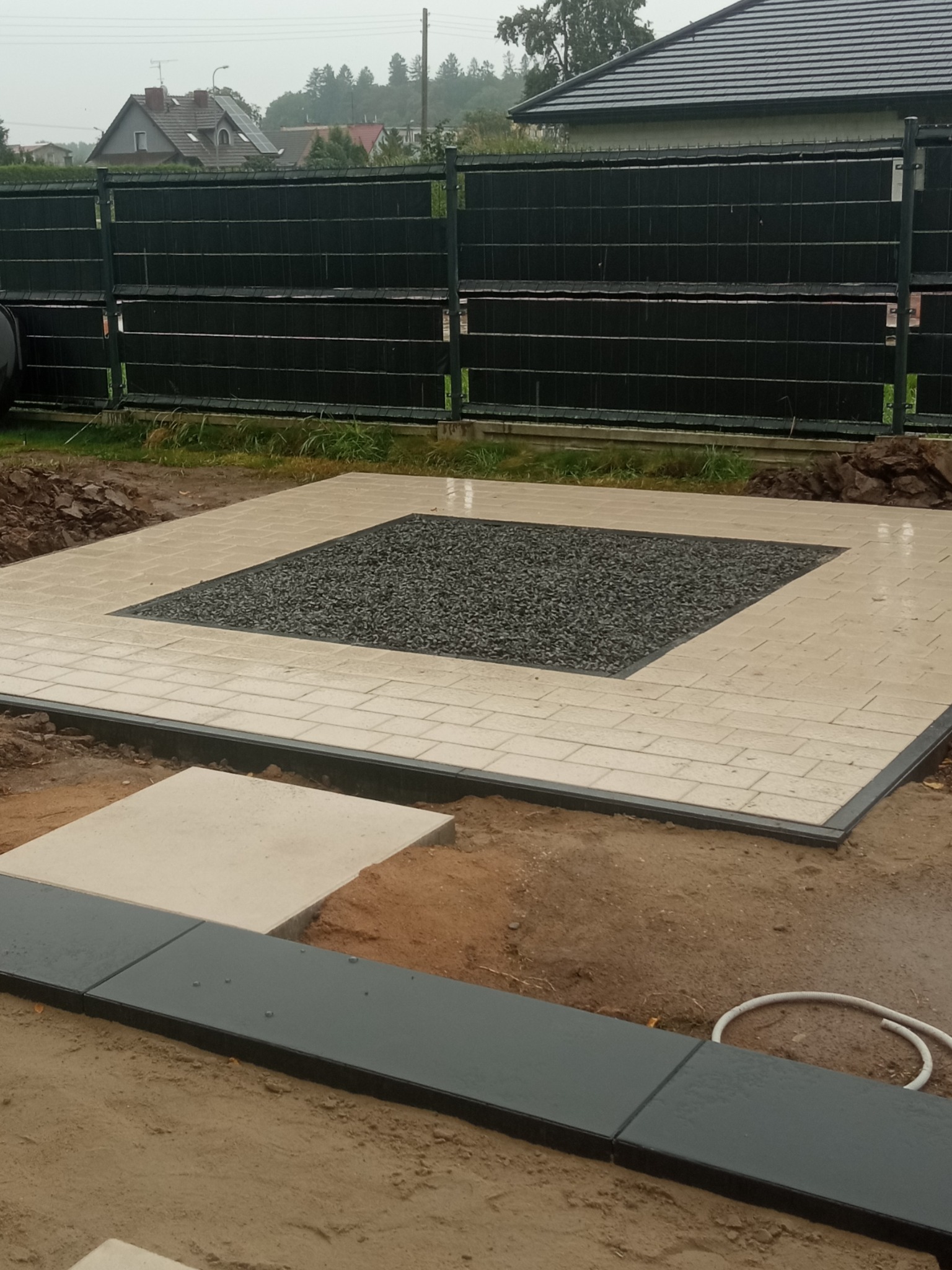Wykończona powierzchnia z jasnej kostki brukowej otaczającej czarne kamienie ozdobne. W tle widoczne ogrodzenie panelowe i zabudowa jednorodzinna.