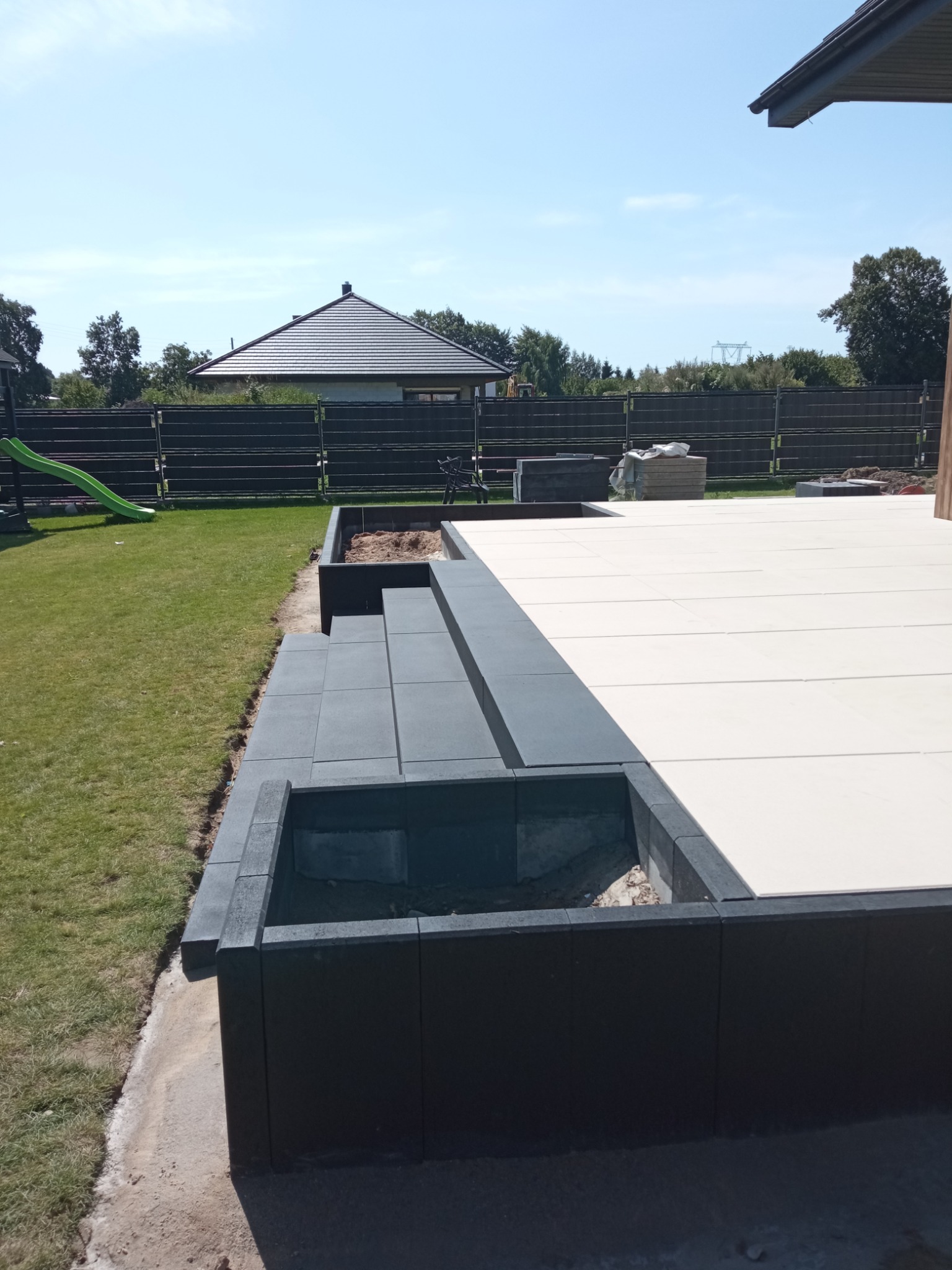 Nowoczesny taras z jasnych płyt i ciemnoszarych obrzeży, z widocznymi schodami i elementami małej architektury ogrodowej w tle, na tle zielonego trawnika.