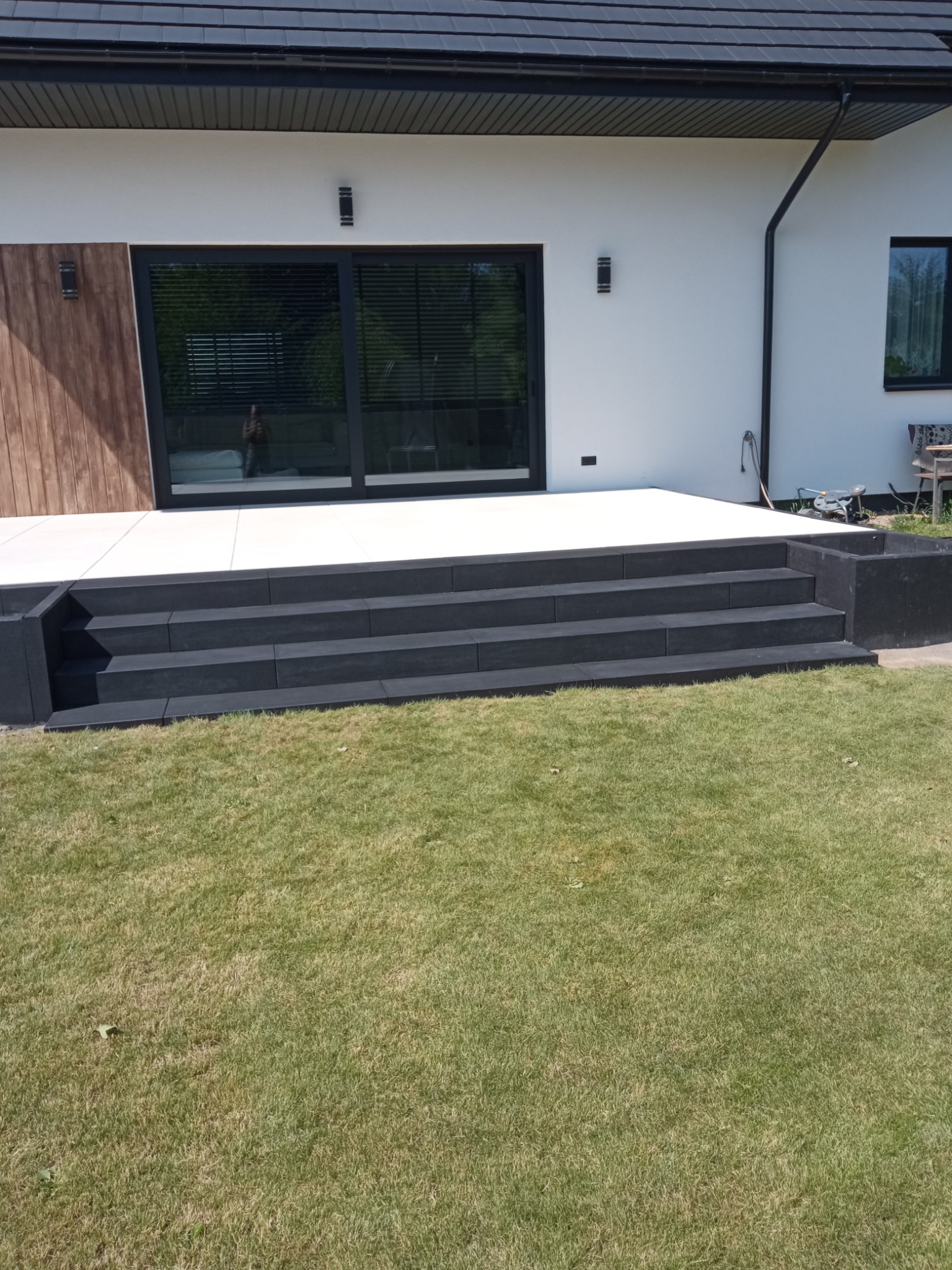 Nowoczesne schody z ciemnej kostki prowadzące na taras z jasnych płyt, przylegające do białego budynku z dużym oknem tarasowym i zadbanym trawnikiem.