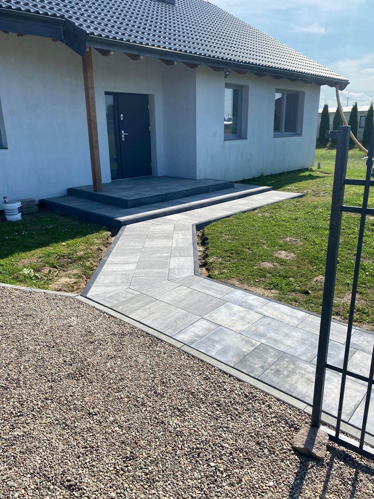 Ułożona kostka brukowa tworzy ścieżkę do domu z szarymi schodami. Obok ścieżki znajduje się obszar wysypany drobnym żwirem i trawnik. W tle widać fragment ogrodzenia.