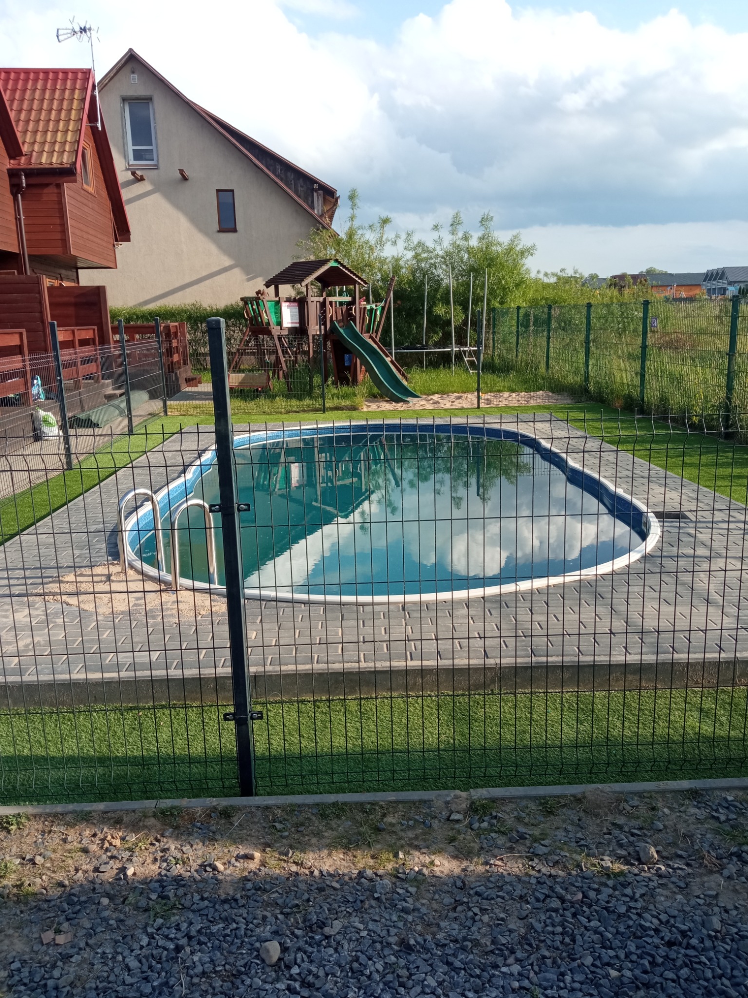 Owalny basen ogrodowy z metalową drabinką, otoczony szarą kostką brukową i zieloną sztuczną trawą, ogrodzony siatką. W tle domy i plac zabaw.