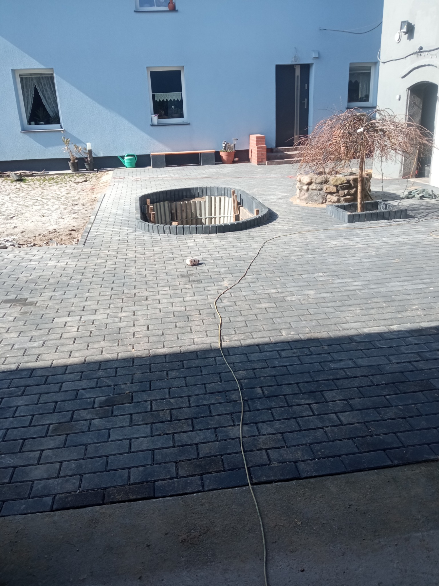 Ułożona kostka brukowa w szarym odcieniu, tworząca ścieżkę wokół wykopu zabezpieczonego obrzeżami. W tle budynek z jasną elewacją i elementami zieleni.