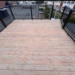 Klaudiusz Wiśniewski - Jasny drewniany taras na dachu budynku z czarną metalową balustradą i widokiem na okolicę. Minimalistyczny design, symetryczne ułożenie desek.