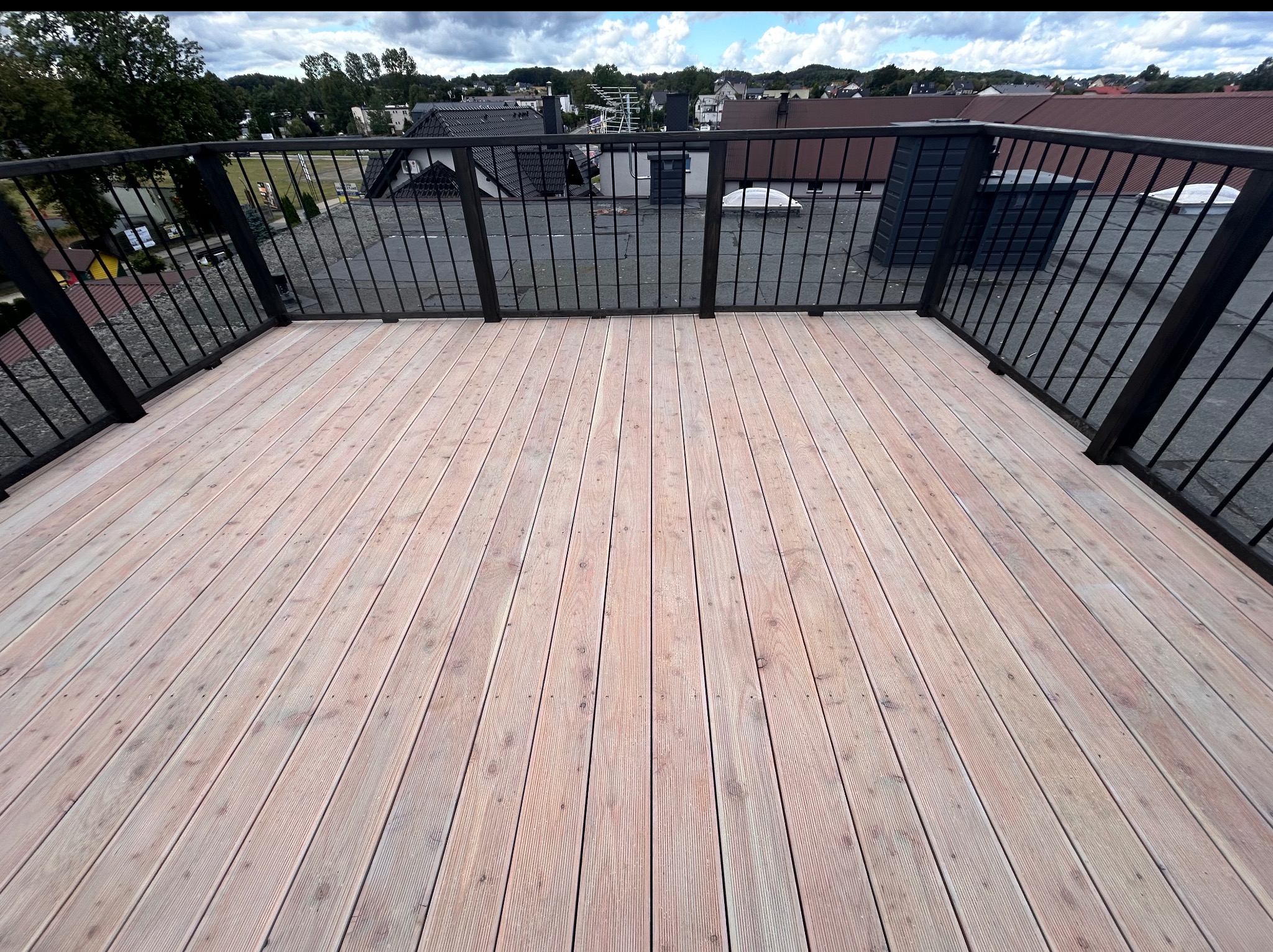 Jasny, drewniany taras na dachu budynku z czarną, metalową balustradą. Widok na okoliczne domy i zieleń w tle, pochmurne niebo.