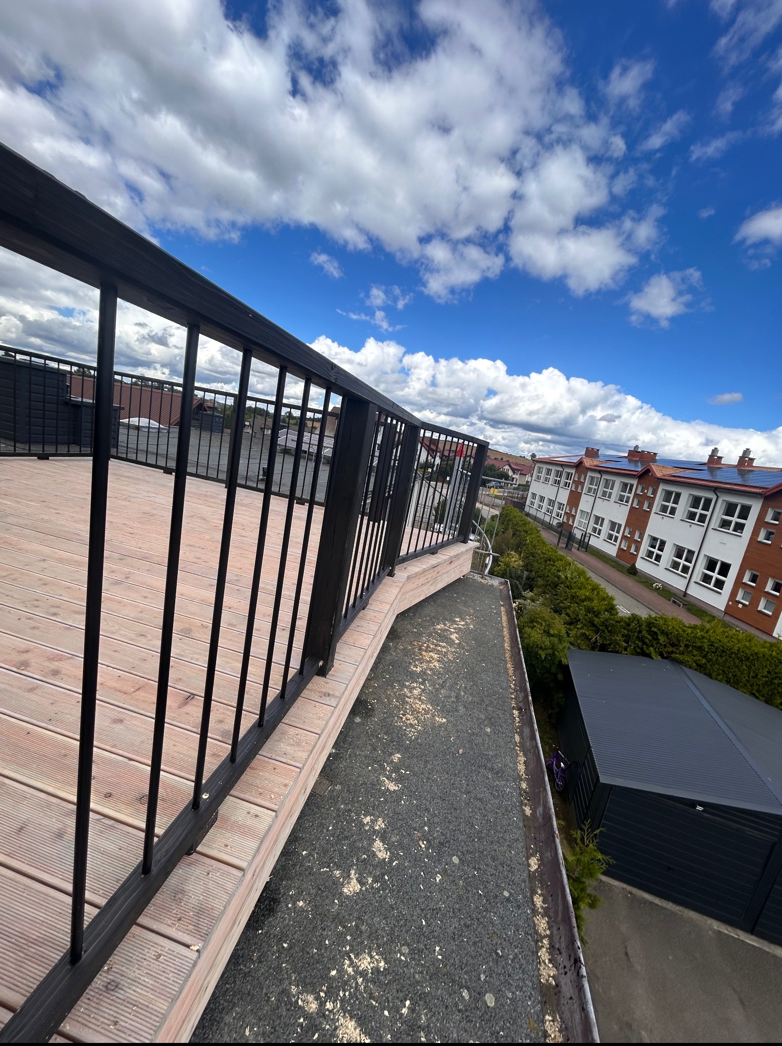 Drewniany taras z czarną, metalową balustradą, widziany z góry, z budynkami w tle i błękitnym niebem z chmurami. Ujęcie z nietypowej perspektywy.