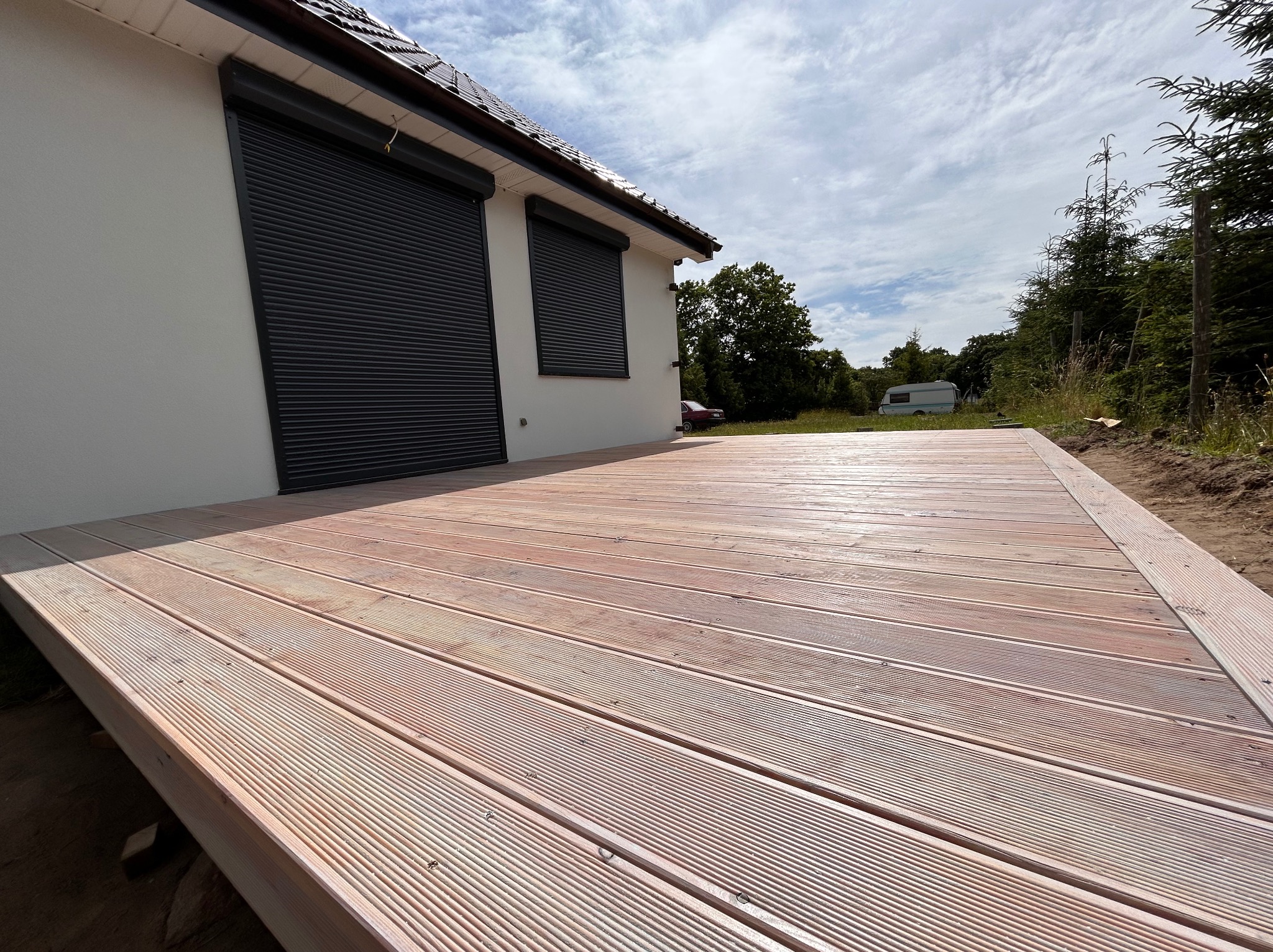 Nowy, jasny taras z desek z ryflowaną powierzchnią, przylegający do białego budynku z szarymi roletami. W tle zieleń drzew i przyczepa kempingowa.