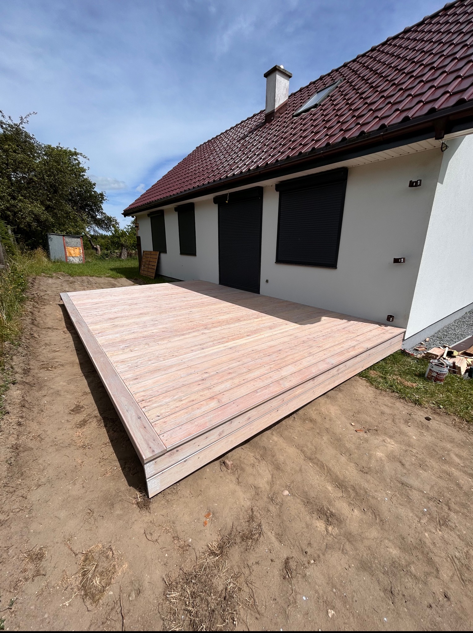 Jasny, drewniany taras przylegający do białego domu z ciemnymi roletami, uchwycony w słoneczny dzień z perspektywy szerokokątnej.