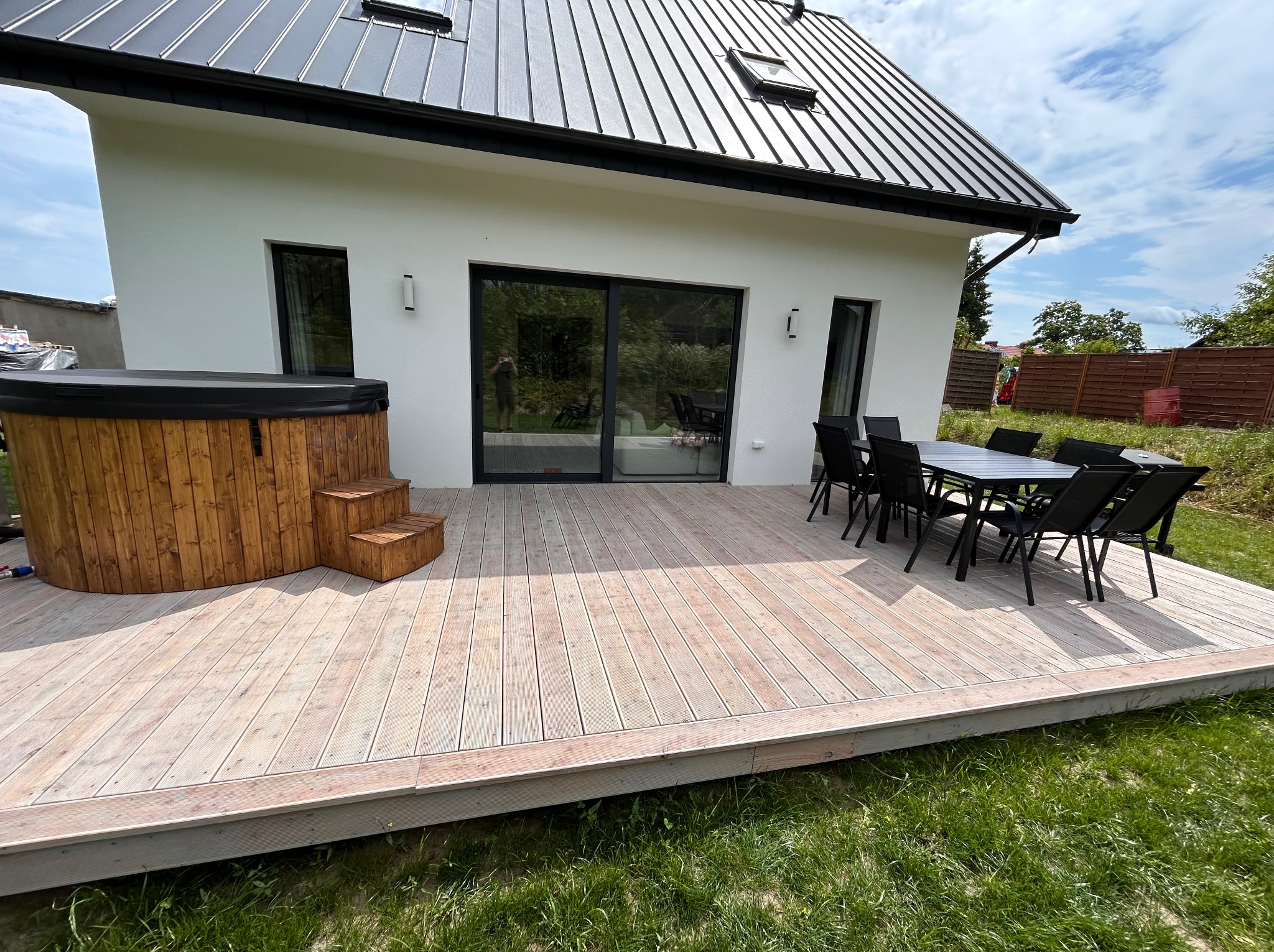 Jasny taras z drewna z jacuzzi i zestawem mebli ogrodowych przy nowoczesnym domu z czarnym dachem. Widok na zewnątrz, trawnik i częściowo ogrodzenie.