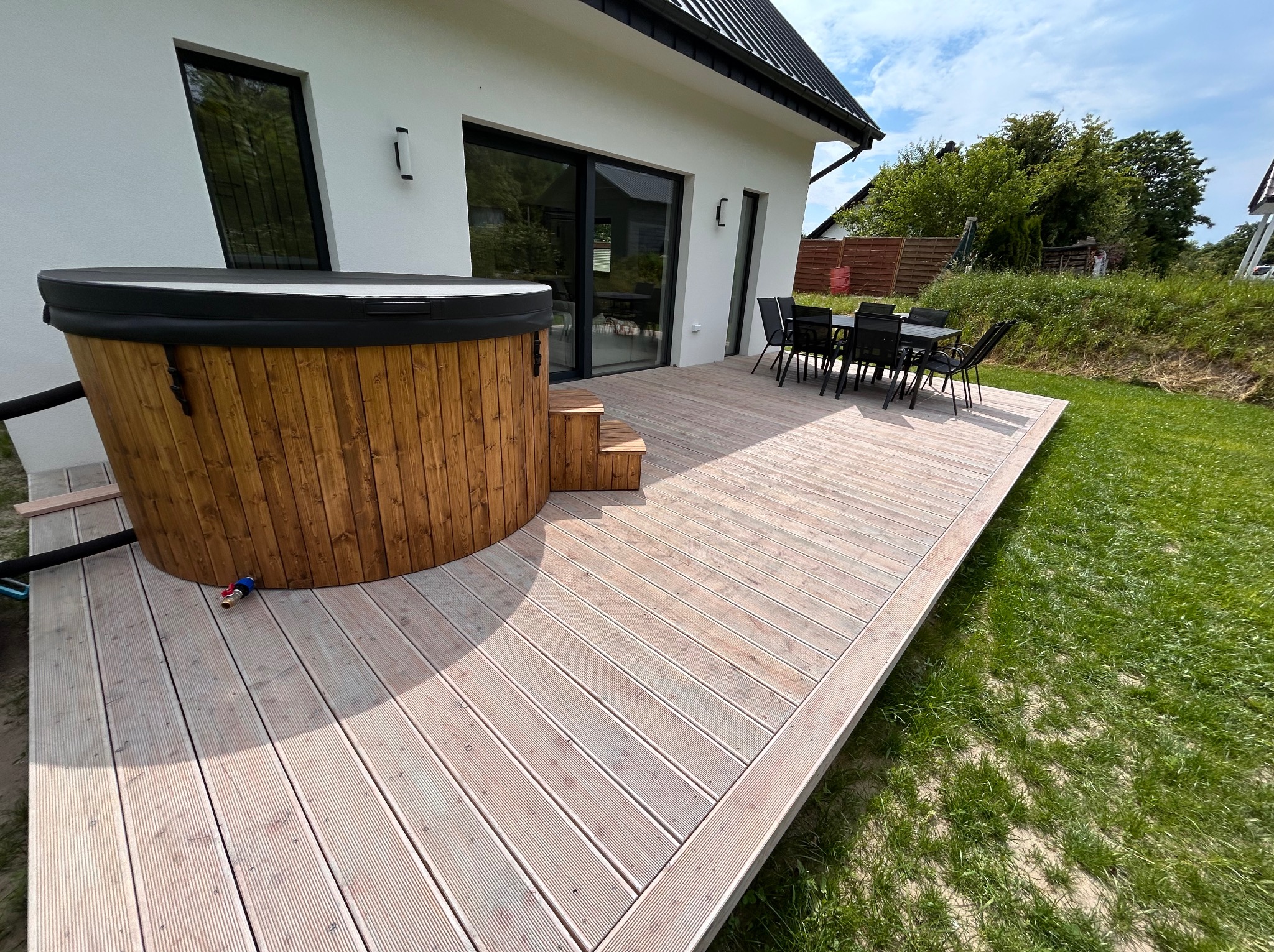 Jasny taras z drewnianą balią i zestawem mebli ogrodowych przy nowoczesnym domu z dużymi oknami. Widok na zielony trawnik i okoliczną zieleń.