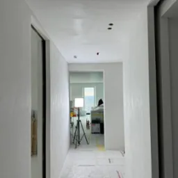 Biały korytarz po gładziowaniu, z widocznymi śladami po szpachlowaniu na ścianach i suficie. Oświetlenie robocze na statywie w tle. Podłoga zabezpieczona folią.