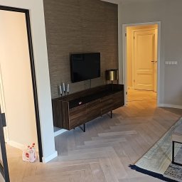 River &.Tree Construction - Salon po remoncie: parkiet w jodełkę, ściana z tapetą w odcieniach brązu, szafka RTV z telewizorem, minimalistyczne świeczniki i lampa. Widok na otwarte drzwi.