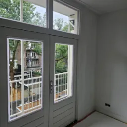 Świeżo pomalowane, białe, dwuskrzydłowe drzwi balkonowe z widokiem na zieleń miejską. Wnętrze po remoncie, z widocznymi gniazdkami elektrycznymi i listwami przypodłogowymi.