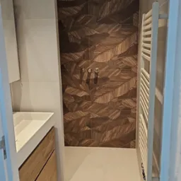 Nowoczesna łazienka z kabiną prysznicową typu walk-in, płytki z motywem liści, umywalka z drewnianą szafką i grzejnik drabinkowy. Minimalistyczny design.