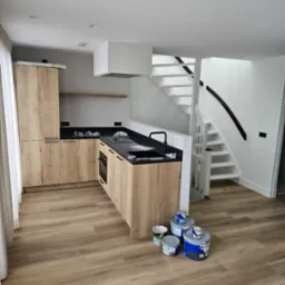 Jasna kuchnia z drewnianymi szafkami i czarnym blatem. Widoczne białe schody i farby malarskie. Podłoga wyłożona panelami imitującymi drewno.