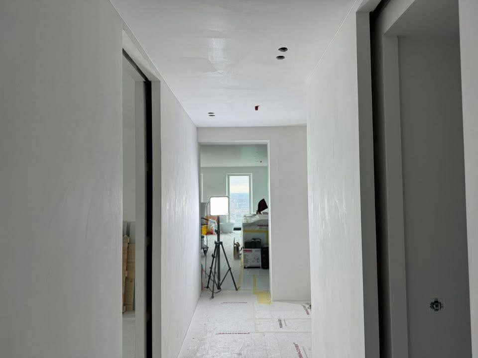 Biały korytarz po gładziowaniu, z widocznymi śladami po szpachlowaniu na ścianach i suficie. Oświetlenie robocze na statywie w tle. Podłoga zabezpieczona folią.