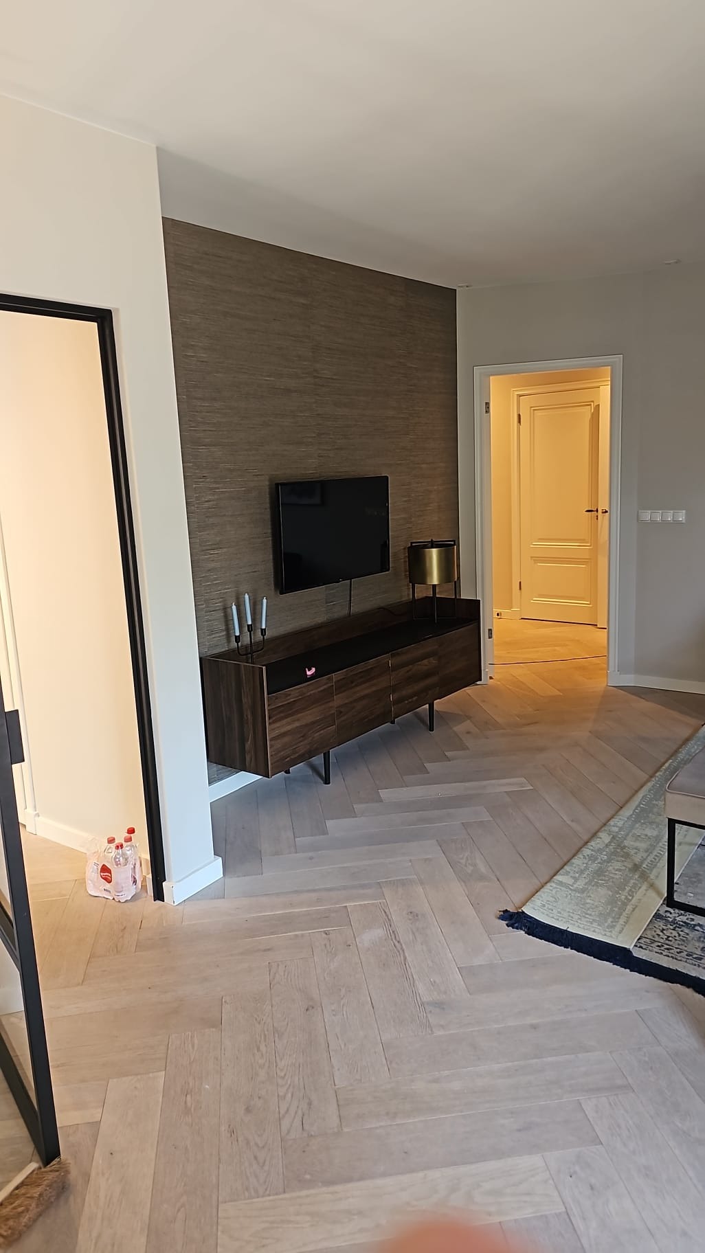 Salon po remoncie: parkiet w jodełkę, ściana z tapetą w odcieniach brązu, szafka RTV z telewizorem, minimalistyczne świeczniki i lampa. Widok na otwarte drzwi.