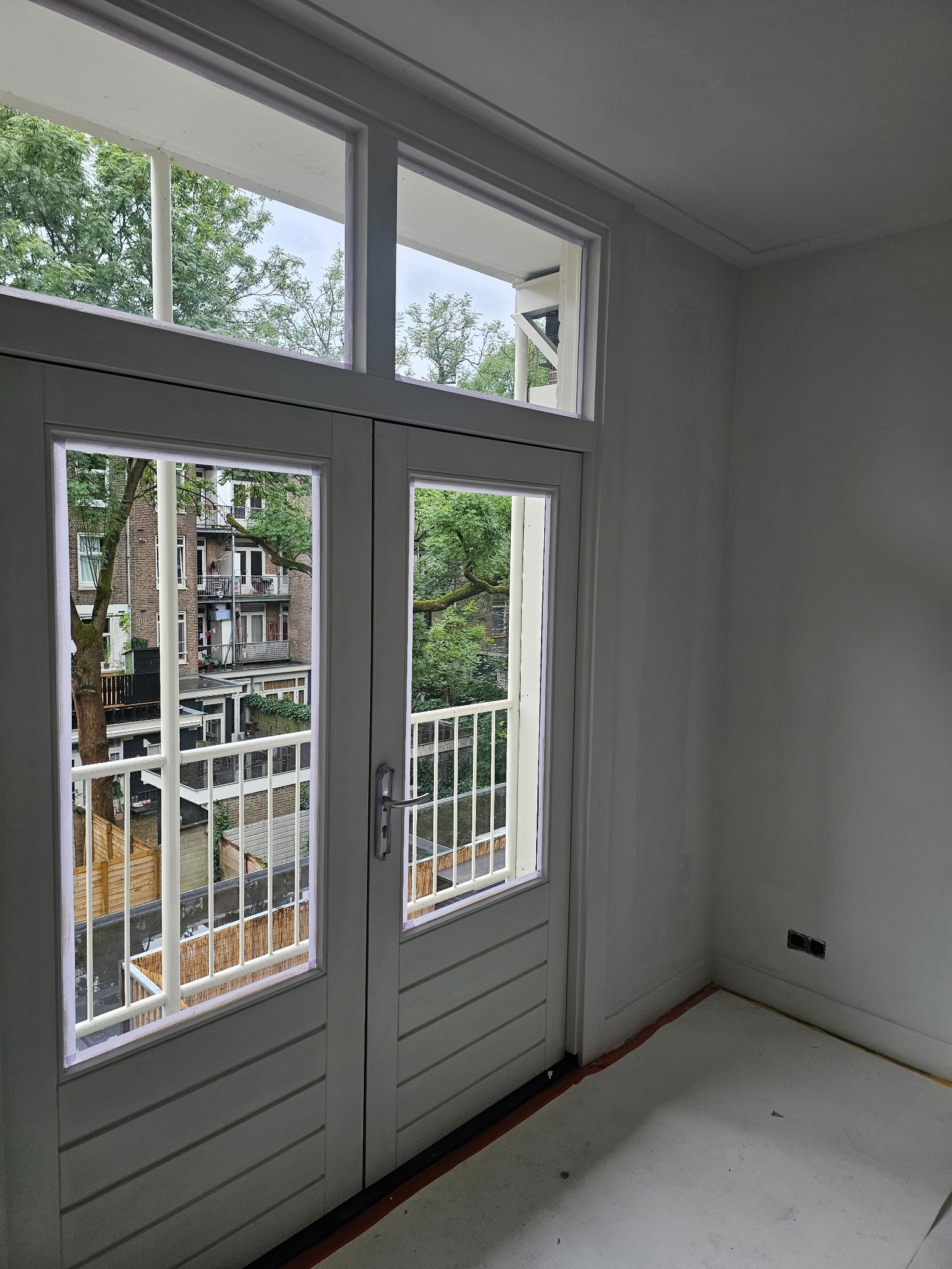 Świeżo pomalowane, białe, dwuskrzydłowe drzwi balkonowe z widokiem na zieleń miejską. Wnętrze po remoncie, z widocznymi gniazdkami elektrycznymi i listwami przypodłogowymi.