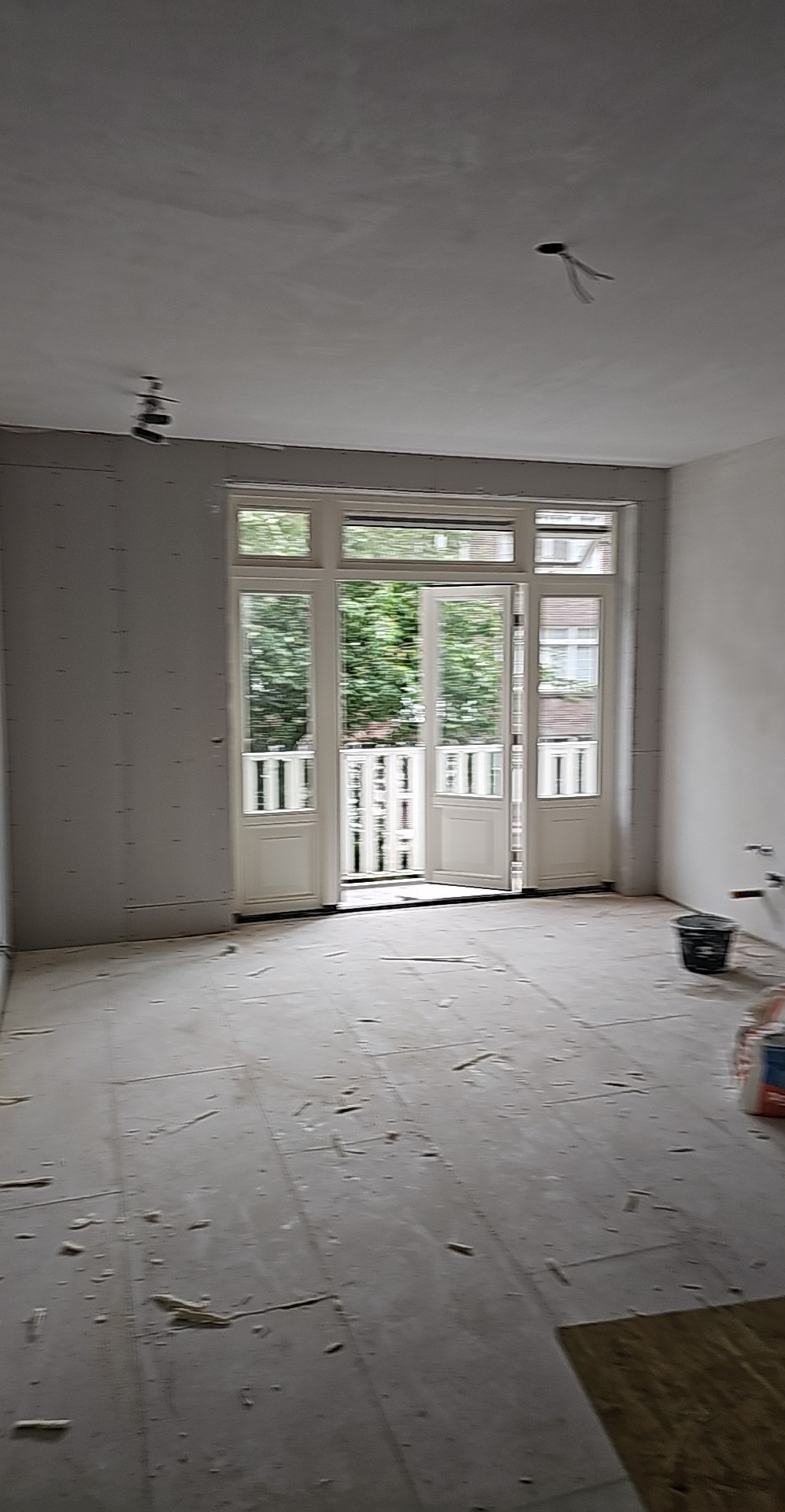 Wnętrze w trakcie remontu: ściany pokryte płytami gipsowo-kartonowymi, przygotowane do szpachlowania. Widoczne otwarte drzwi balkonowe i resztki materiałów na podłodze.