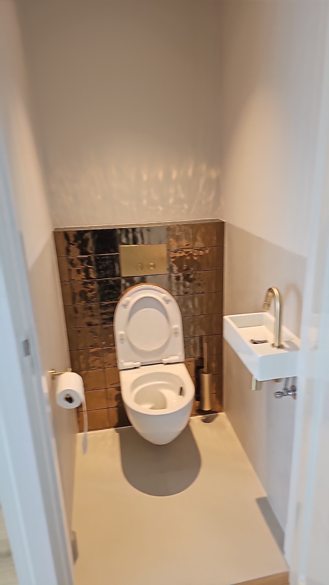 Nowoczesna łazienka: biała toaleta z deską w kolorze drewna, złote płytki na ścianie, biała umywalka ze złotą armaturą. Minimalistyczny design z akcentami złota.