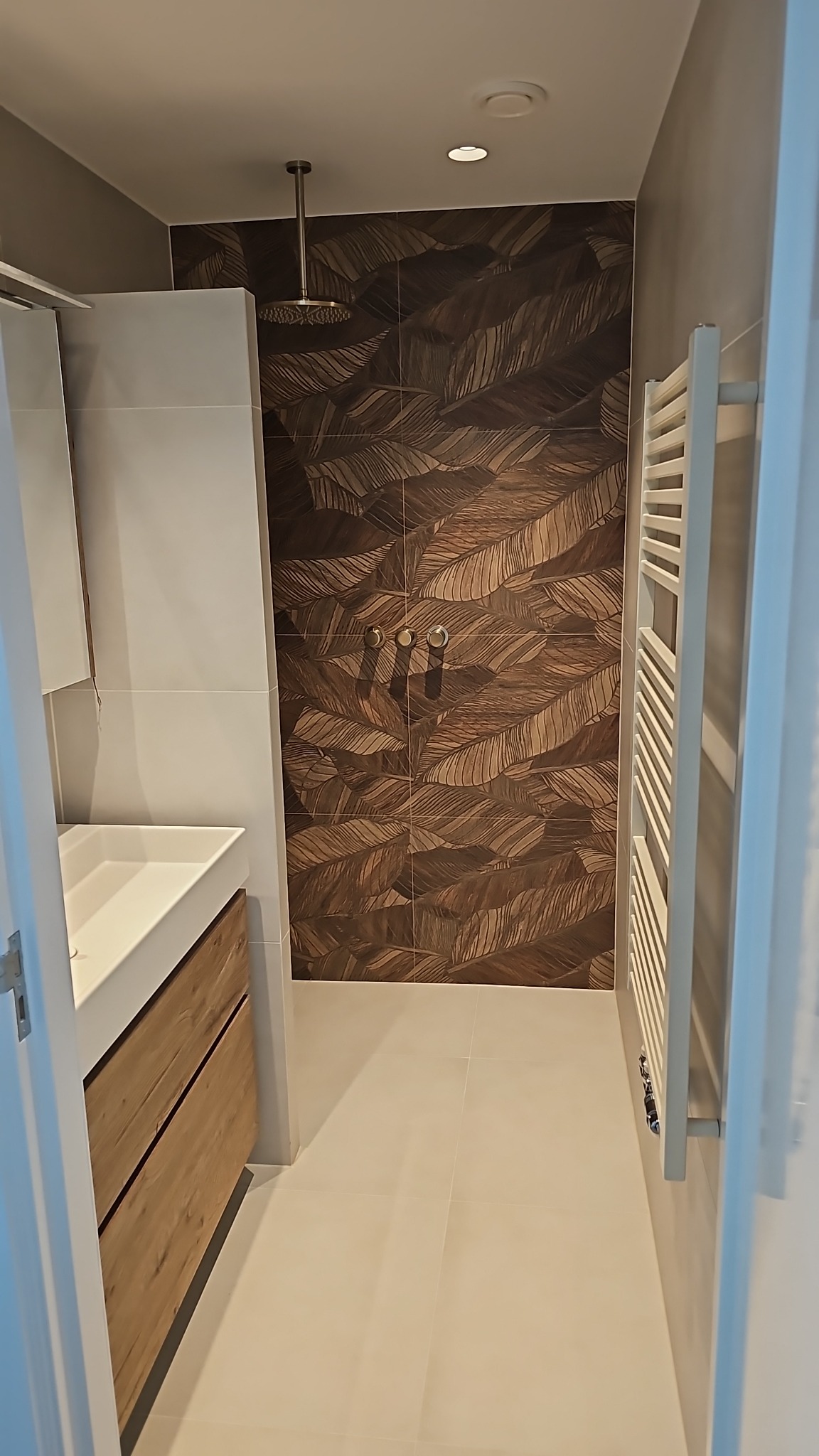 Nowoczesna łazienka z kabiną prysznicową typu walk-in, płytki z motywem liści, umywalka z drewnianą szafką i grzejnik drabinkowy. Minimalistyczny design.