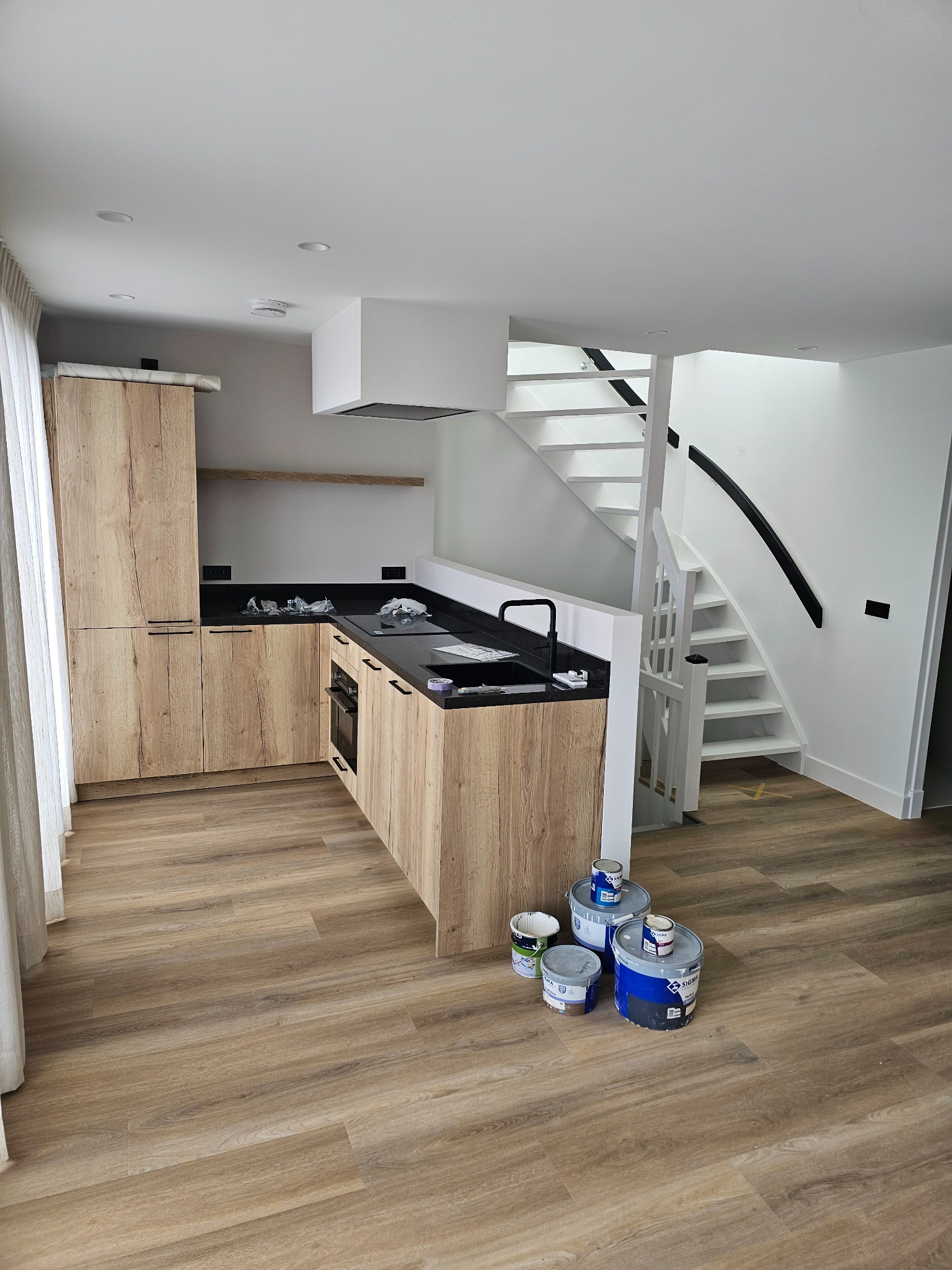 Jasna kuchnia z drewnianymi szafkami i czarnym blatem. Widoczne białe schody i farby malarskie. Podłoga wyłożona panelami imitującymi drewno.