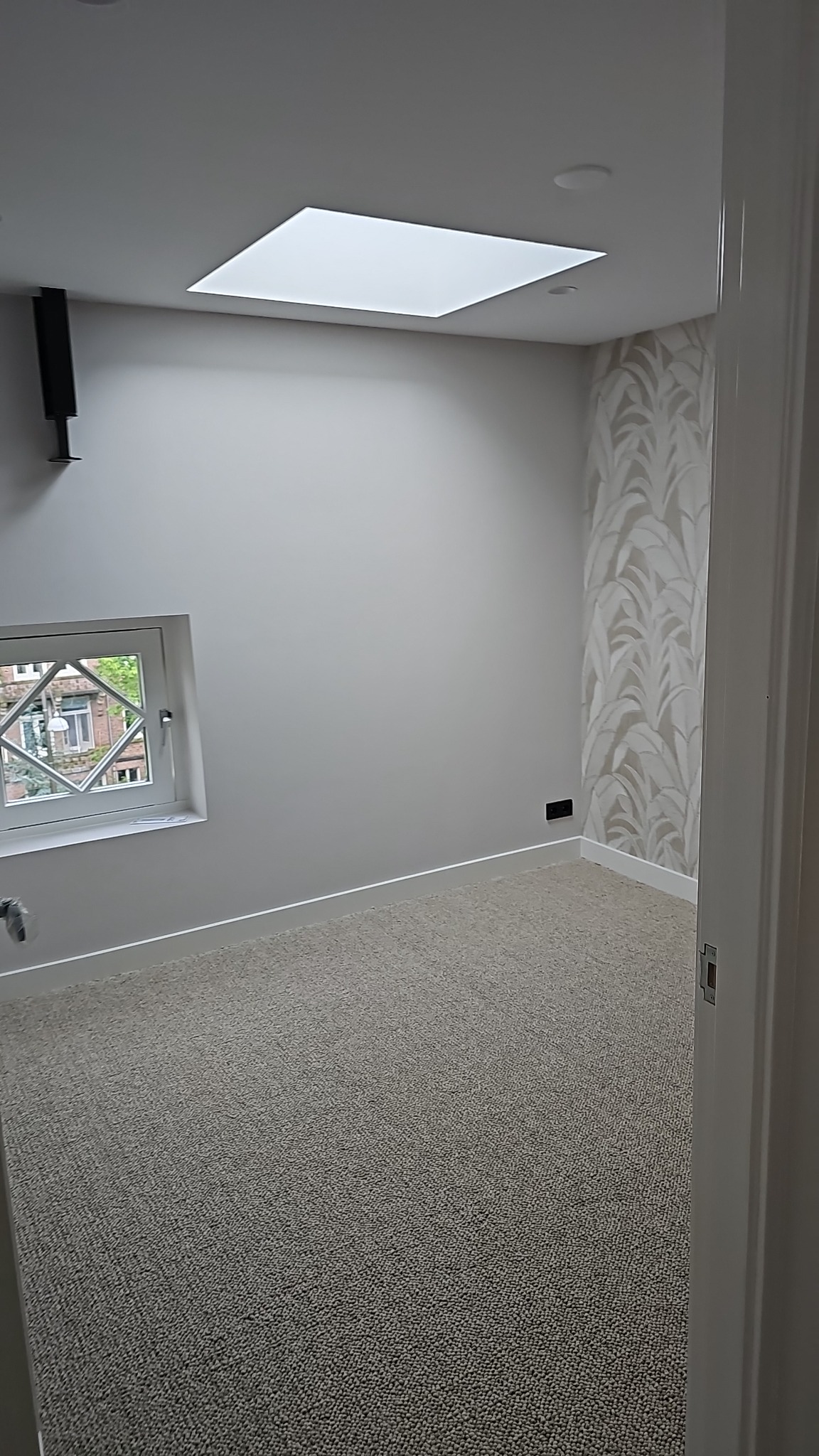 Wykończone wnętrze pokoju po remoncie: szare ściany, okno z ozdobną kratą, wzorzysta tapeta na jednej ścianie, dywan i sufit z oknem dachowym. Widok zza otwartych drzwi.