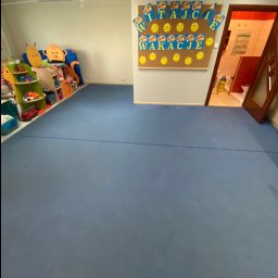 Clean It Firma Sprzątająca Specialistyczne Pranie Tapiceki - Pusty pokój dziecięcy z niebieską podłogą, kolorowymi dekoracjami i tablicą z napisem 'Witajcie Wakacje'. Widok na salę zabaw i toaletę z otwartymi drzwiami.