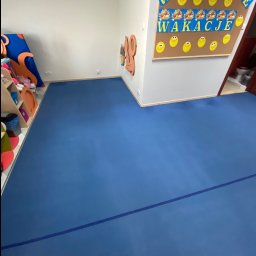 Clean It Firma Sprzątająca Specialistyczne Pranie Tapiceki - Błękitna podłoga z linią w sali przedszkolnej, dekoracje na ścianach i regał z zabawkami. Widok na toaletę w tle. Czysta przestrzeń po sprzątaniu.