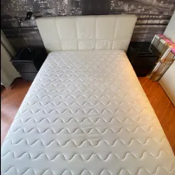 Biały materac na łóżku z pikowanym zagłówkiem, widok z góry. W tle nocny pejzaż miasta na fototapecie i czarne szafki nocne z lampką.