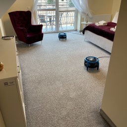 Clean It Firma Sprzątająca Specialistyczne Pranie Tapiceki - Pokój hotelowy z szarym dywanem suszonym przez dwa niebieskie wentylatory po czyszczeniu. W tle balkon z widokiem na góry, fotel i łóżko.