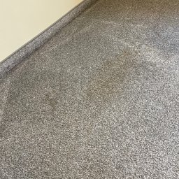 Clean It Firma Sprzątająca Specialistyczne Pranie Tapiceki - Szary dywan z krótkim włosiem, ułożony w rogu pokoju, widoczny fragment ściany w odcieniu kremowym i szara listwa przypodłogowa. Tekstura dywanu jest wyraźna.