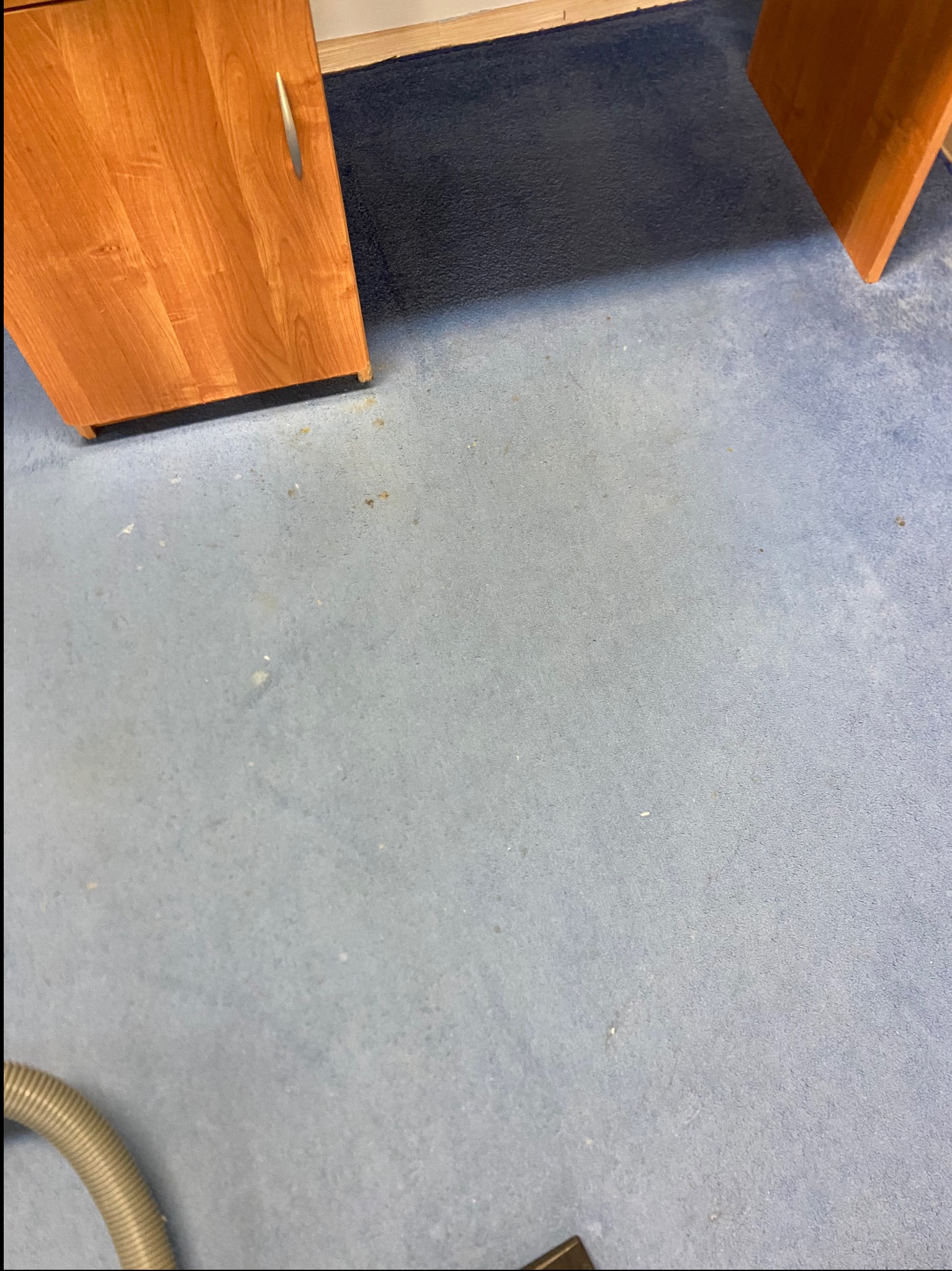 Niebieski dywan w biurze z widocznymi plamami, częściowo widoczny odkurzacz i meble biurowe z drewna. Widok z góry, plamy na dywanie.