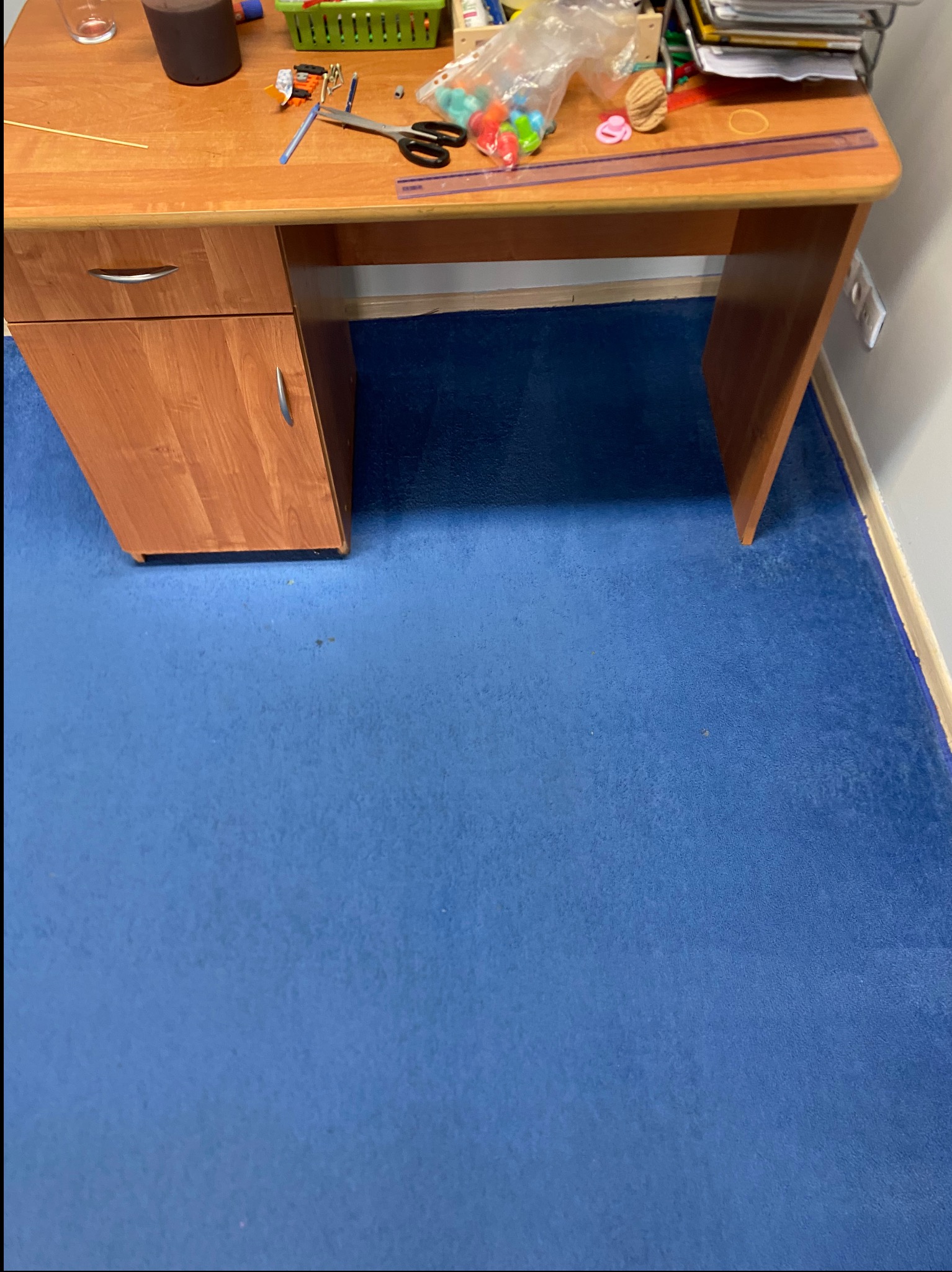 Niebieska wykładzina dywanowa pod drewnianym biurkiem z szufladami, na którym leżą nożyczki, długopis, linijka i inne przedmioty.