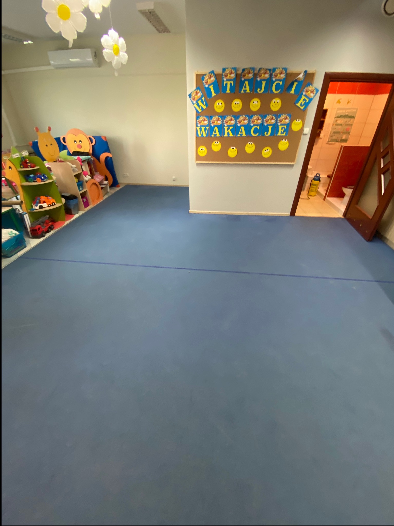 Pusty pokój dziecięcy z niebieską podłogą, kolorowymi dekoracjami i tablicą z napisem 'Witajcie Wakacje'. Widok na salę zabaw i toaletę z otwartymi drzwiami.