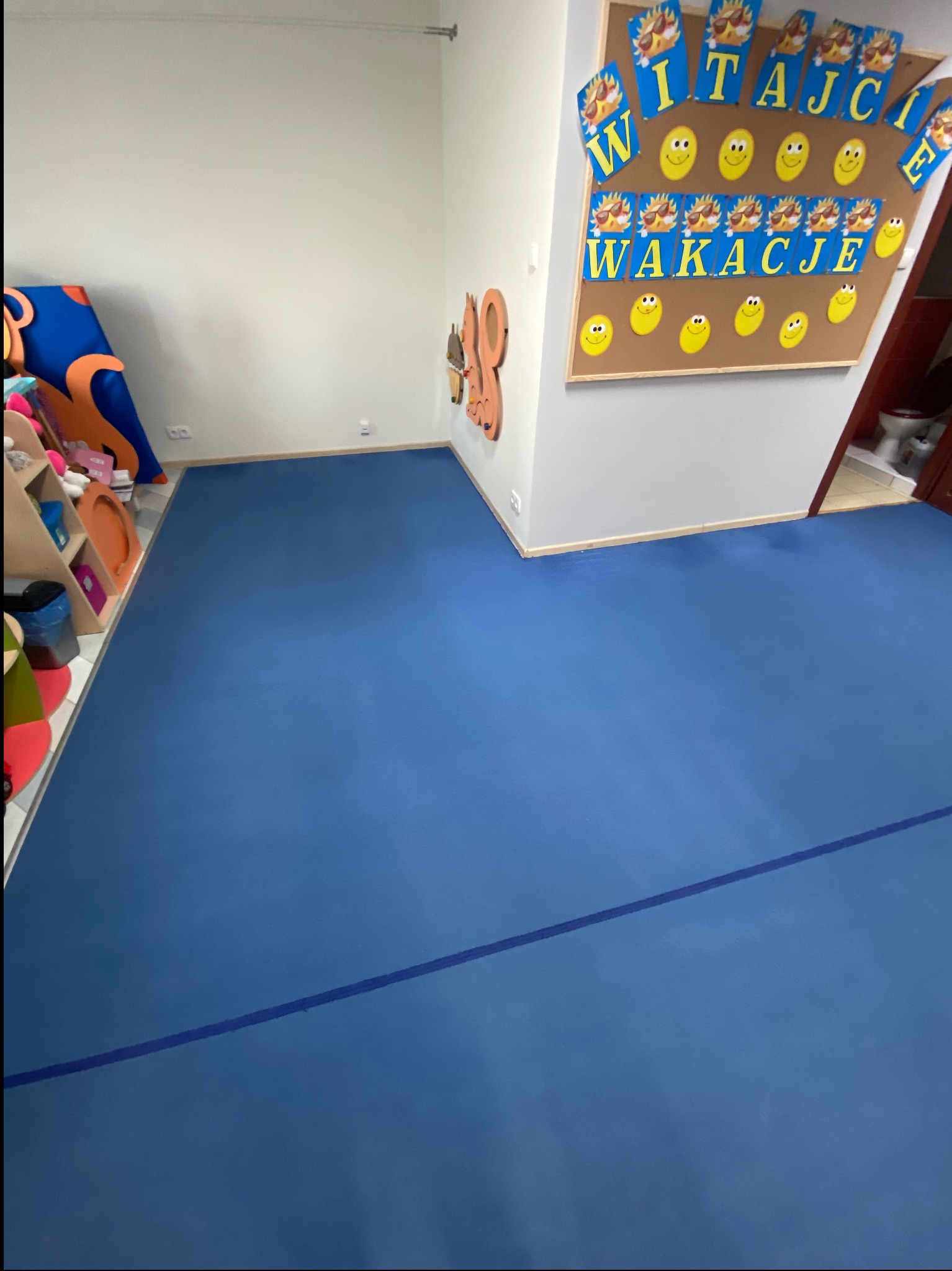 Błękitna podłoga z linią w sali przedszkolnej, dekoracje na ścianach i regał z zabawkami. Widok na toaletę w tle. Czysta przestrzeń po sprzątaniu.