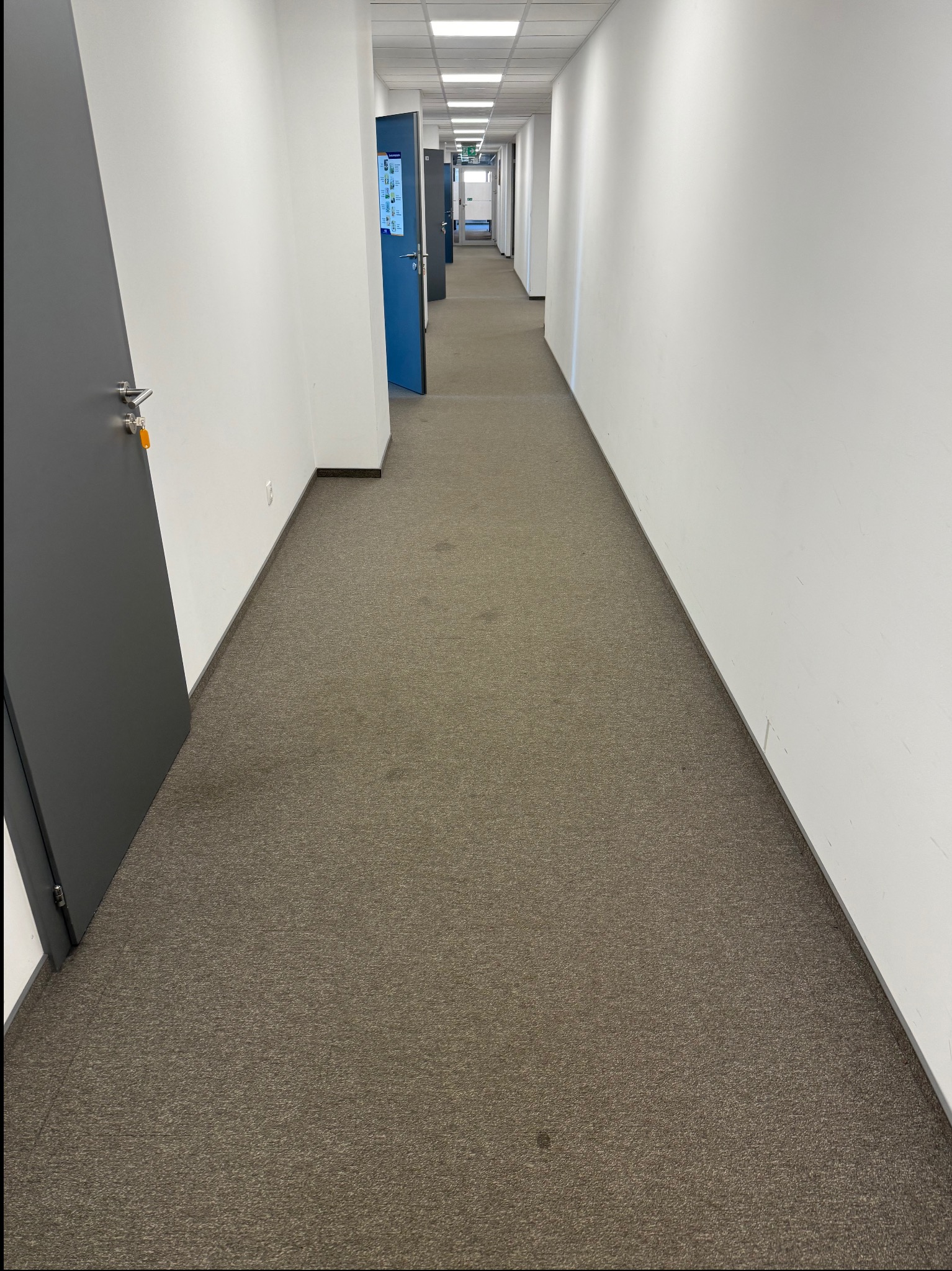 Długi korytarz biurowy z brązową wykładziną i białymi ścianami. Widoczne drzwi po lewej stronie i w oddali. Światło sufitowe równomiernie oświetla przestrzeń.